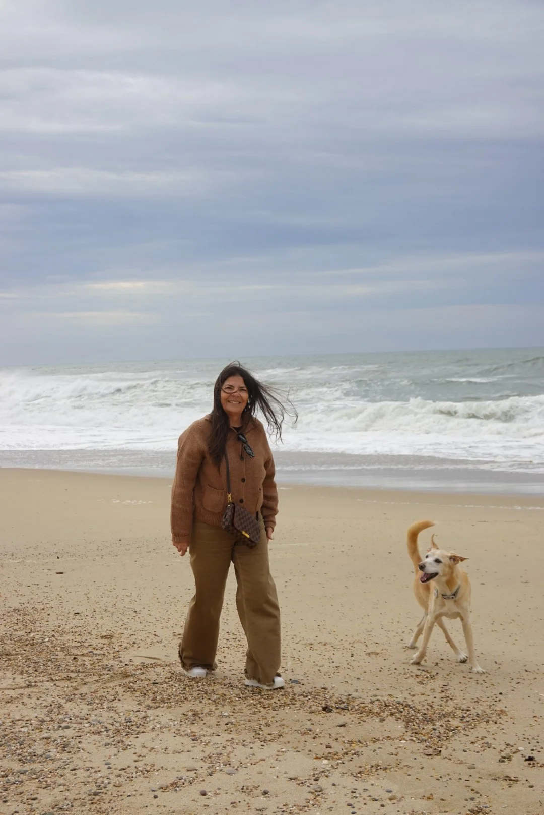 Eine Frau und ein Hund am Strand bei bedecktem Himmel, sie lachen und genießen die Meeresküste.