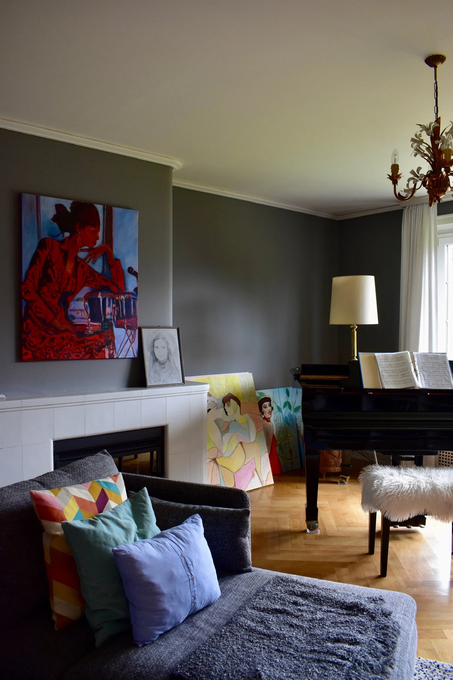 Ein Wohnzimmer mit dunklem Sofa, bunten Kissen, einem Kamin, bunten Gemälden, einer Stehlampe, einem Klavier mit Notenbüchern und weißen Vorhängen bei Tageslicht.