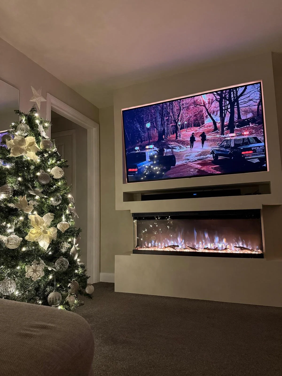 A living room decorated for Christmas with a lit Christmas tree decorated with ornaments and lights on the left, a mounted TV showing a scene from film above a modern electric fireplace with flames within a media wall with a door in the background.