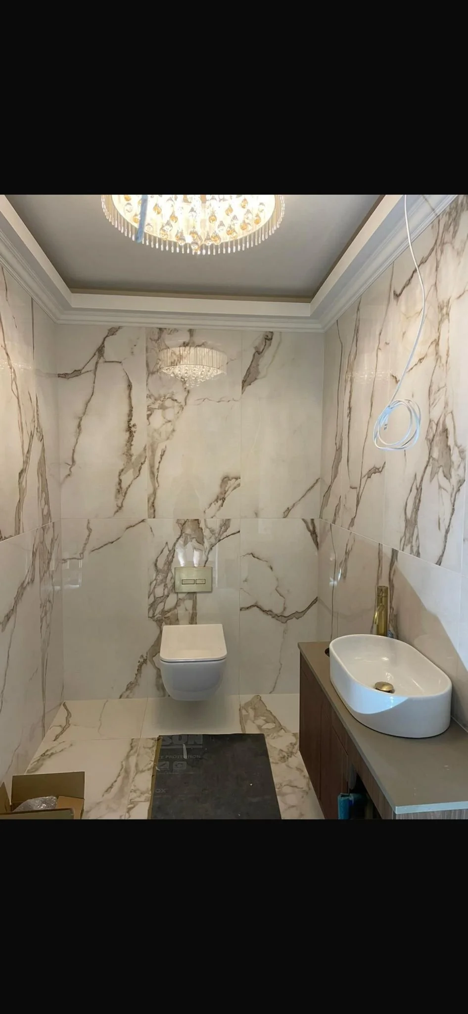 A bathroom with marble walls, a wall-mounted toilet, a small sink with a wooden cabinet, a chandelier lighting fixture on the ceiling.