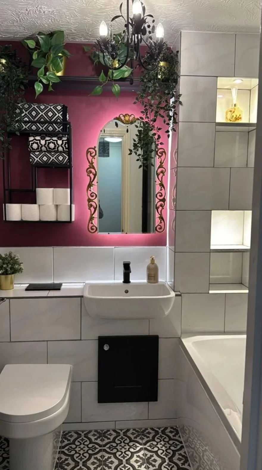 Bathroom with a pink accent wall, a mirror with gold details, a small white sink, a toilet, and a bathtub. Green plants and a chandelier hang from the ceiling, and there are decorative items on shelves.