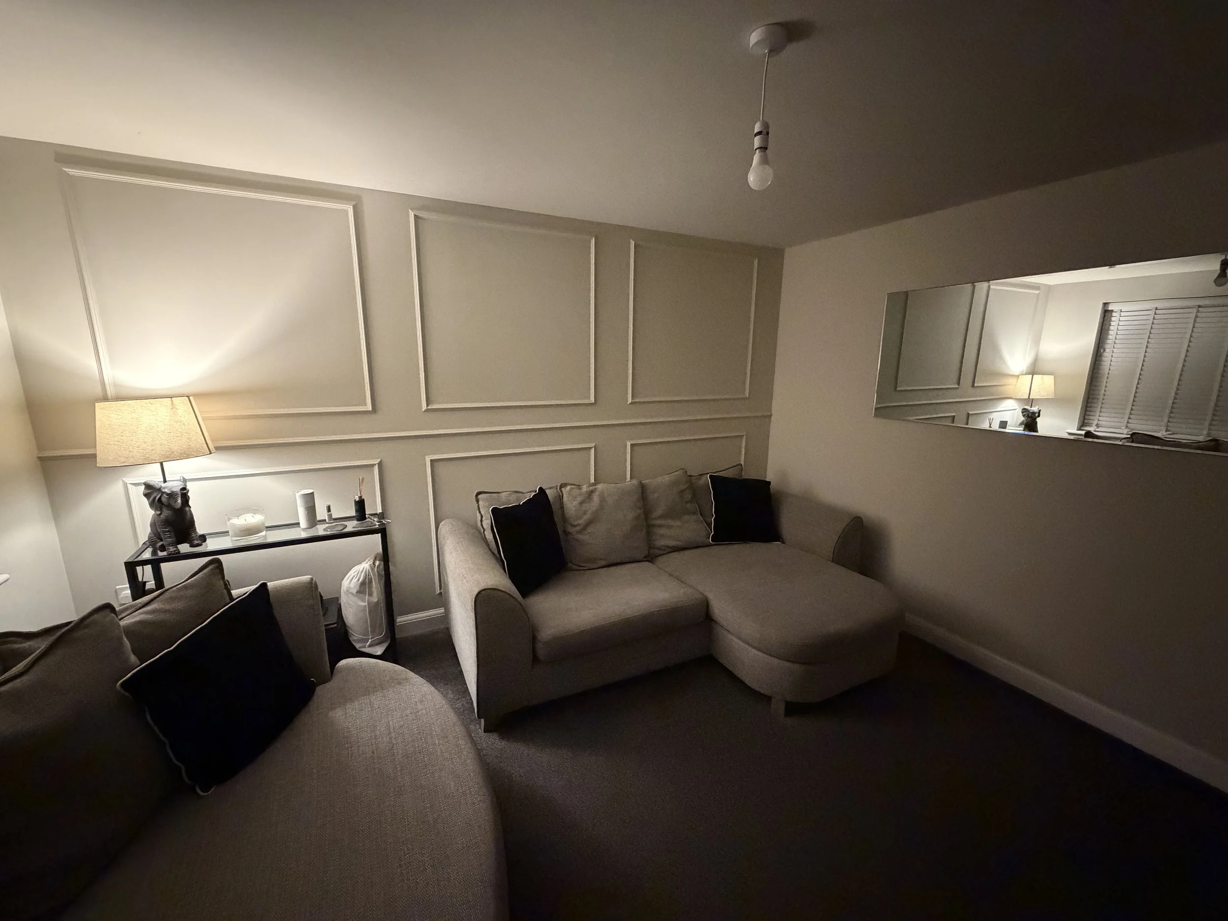 Living room with beige sectional sofa, dark cushions, wall decor, a side table with a lamp, a statue, and a mirror reflecting another lamp and window with blinds.