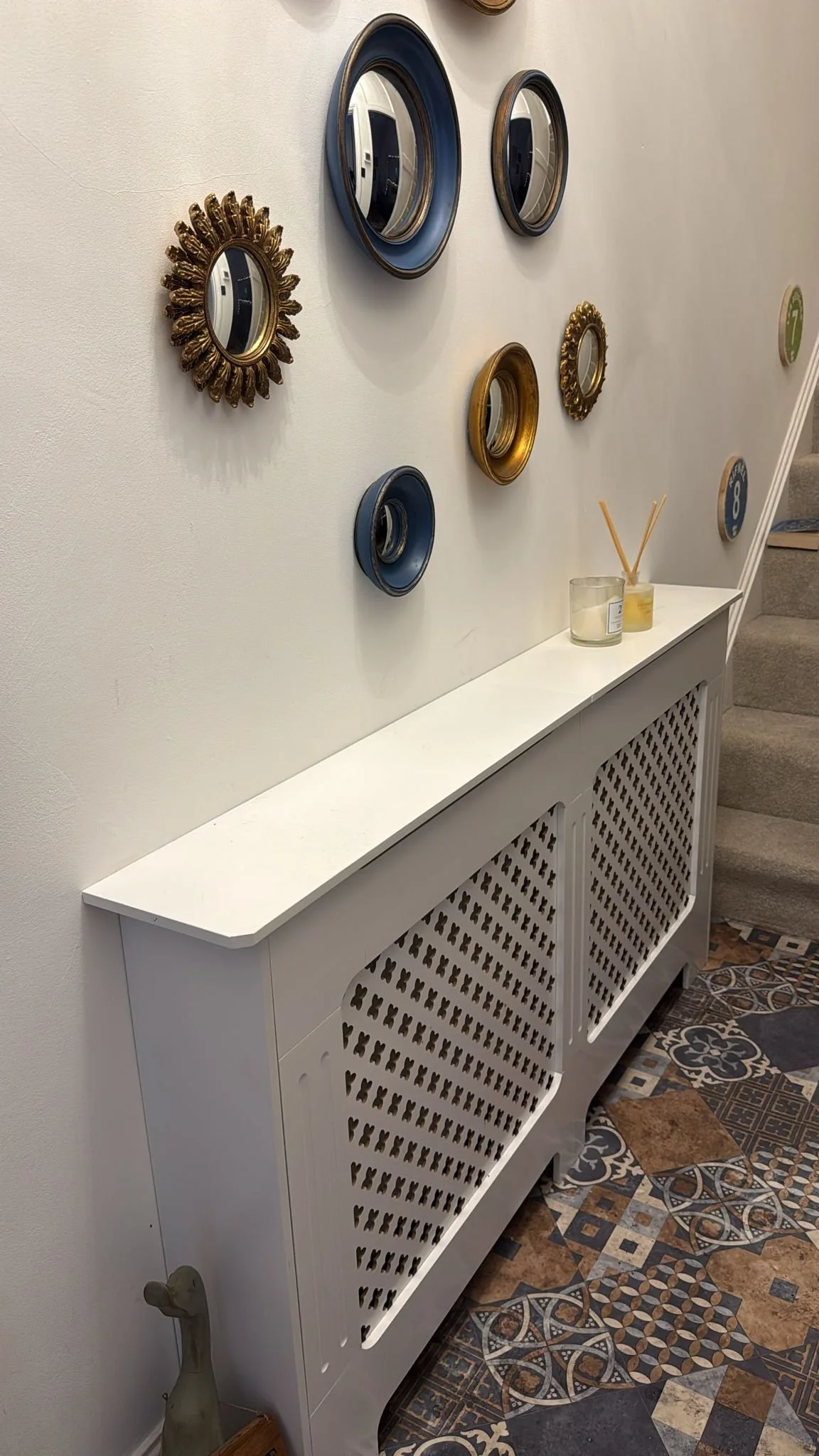 Decorative wall with various vintage-style mirrors and a white radiator cover below, placed in a room with patterned tile flooring.