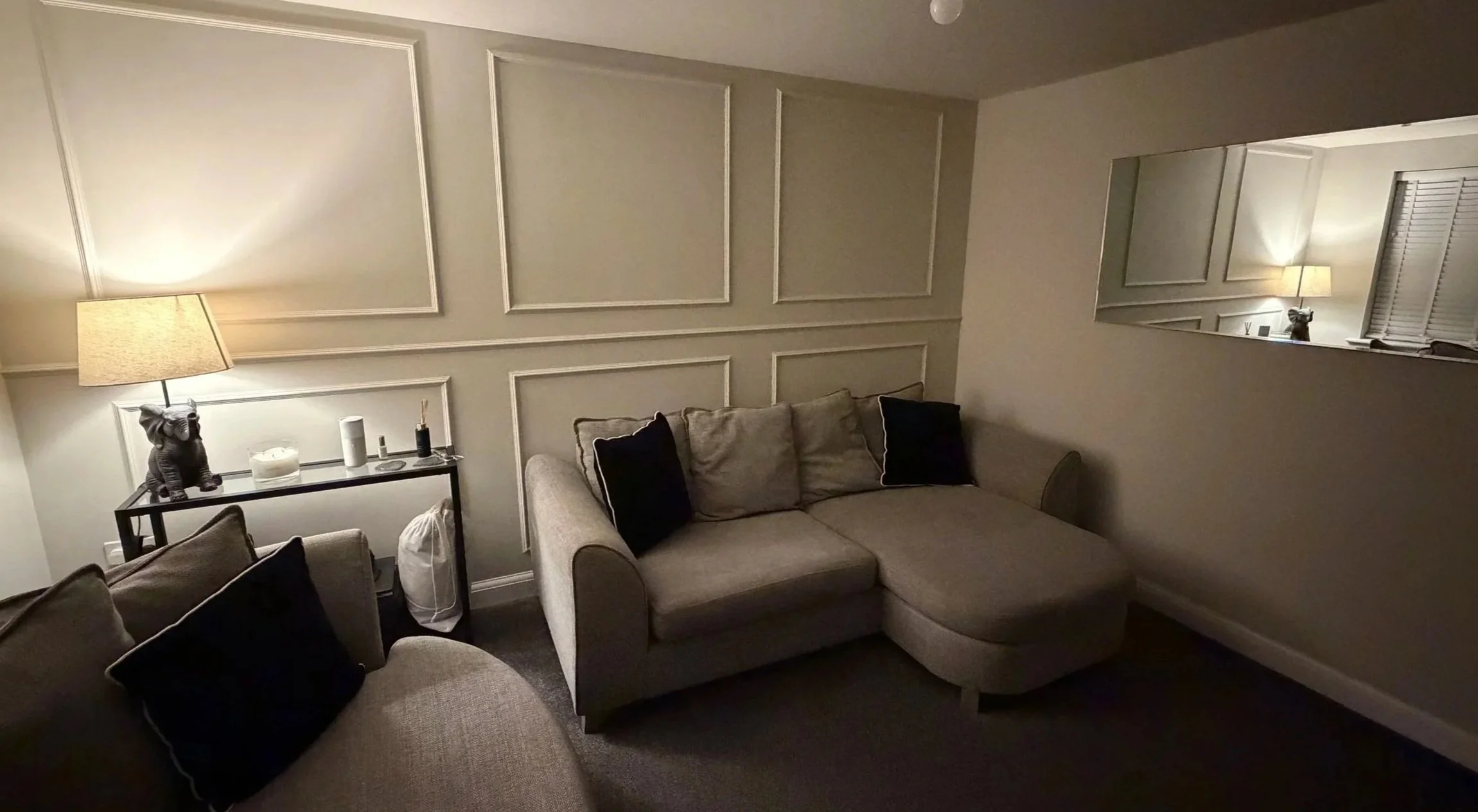 A cozy living room with beige couches, black pillows, a side table with a lamp, a decorative elephant figurine, and a mirror reflecting the room's lighting.