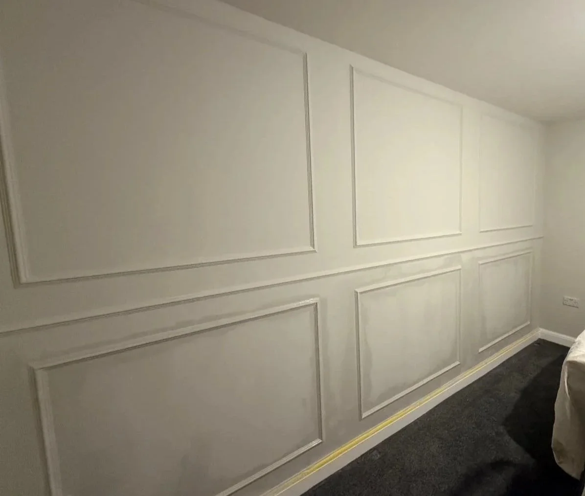 White wall with decorative molding panels in a room, dark carpeted floor, and partially visible bed with white bedding.