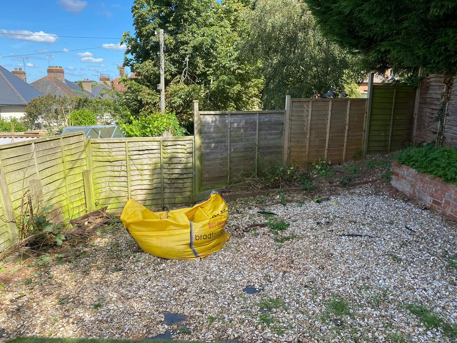 A backyard garden with a graveled ground, partly fenced with wooden panels, some of which are aged and weathered. There is a yellow bag, possibly for garden waste, on the ground near the fence. In the background, there are tall trees and neighboring houses with chimneys under a blue sky with a few clouds.