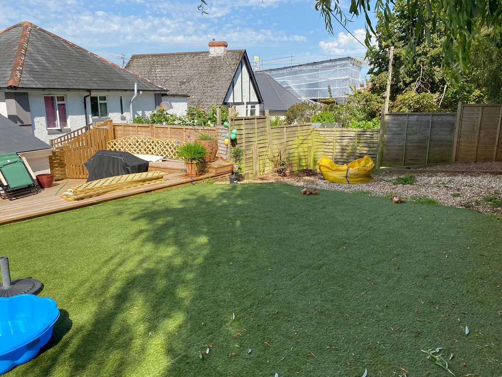 A backyard with a grassy lawn, a small deck with a grill and lawn chair, and a wooden fence. There are trees casting shadows and some outdoor toys and decorations.