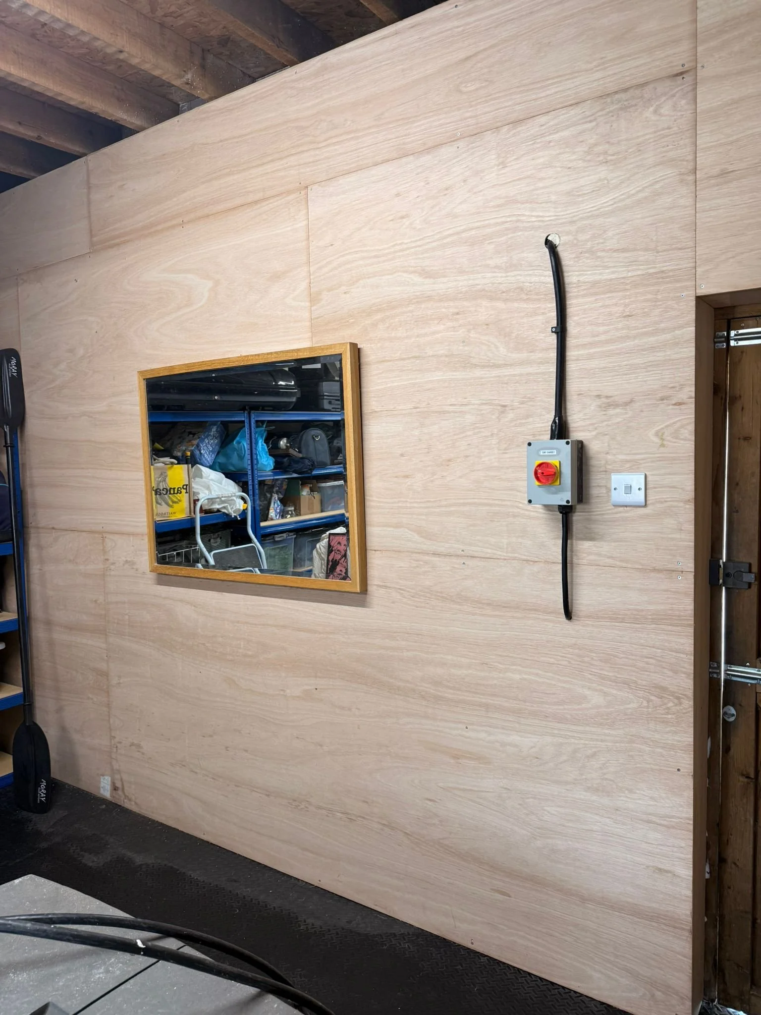 A large ply-lined garage wall with a mirror, a light switch, and a rotary isolator with black cable for an EV charger.