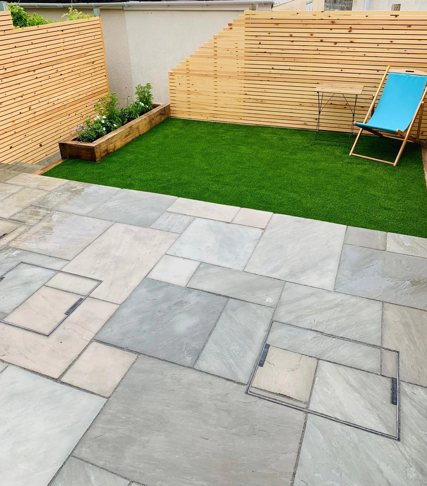 A backyard with a stone patio, a small patch of artificial grass, a wooden fence, a blue lounge chair, a small table, and a flower bed with plants.