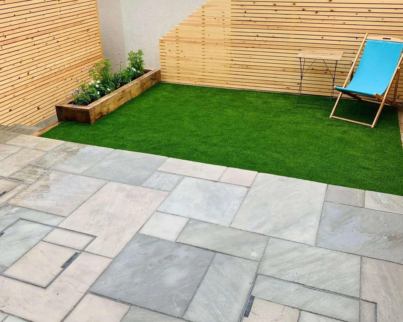 Small backyard patio with stone tiles, a grass patch, a wooden planter with flowers, a wooden privacy fence, a blue outdoor lounge chair, and a small side table.
