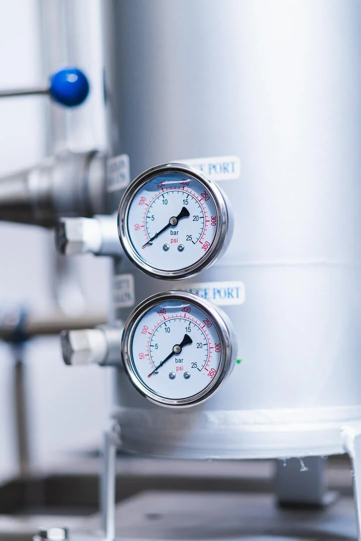 Close-up of two pressure gauges on a white tank, measuring in bar and psi, with pipes attached.