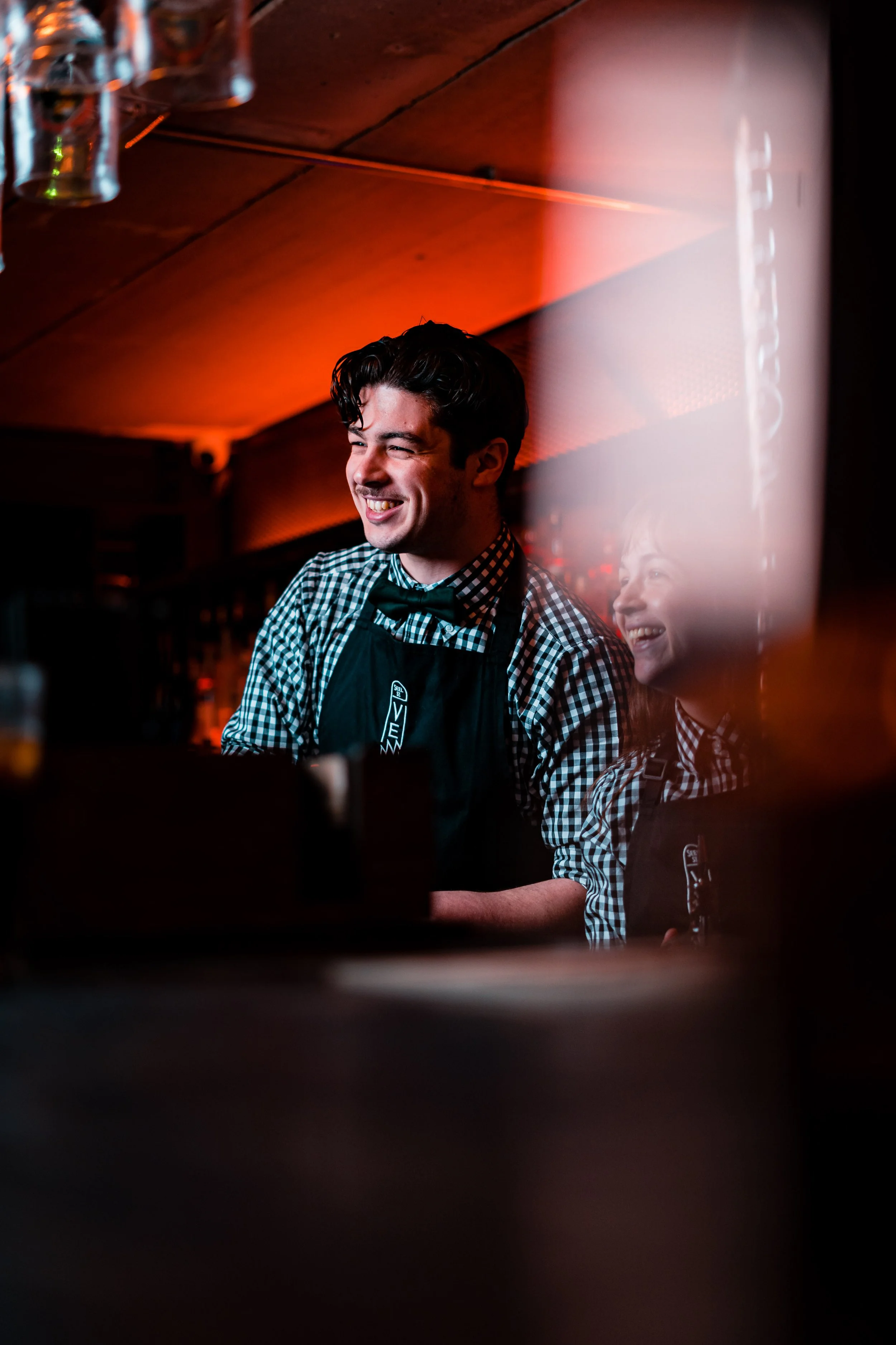 A young male bartender wearing a checkered shirt, black bow tie, and apron, smiling while working behind a bar in a dimly lit setting.