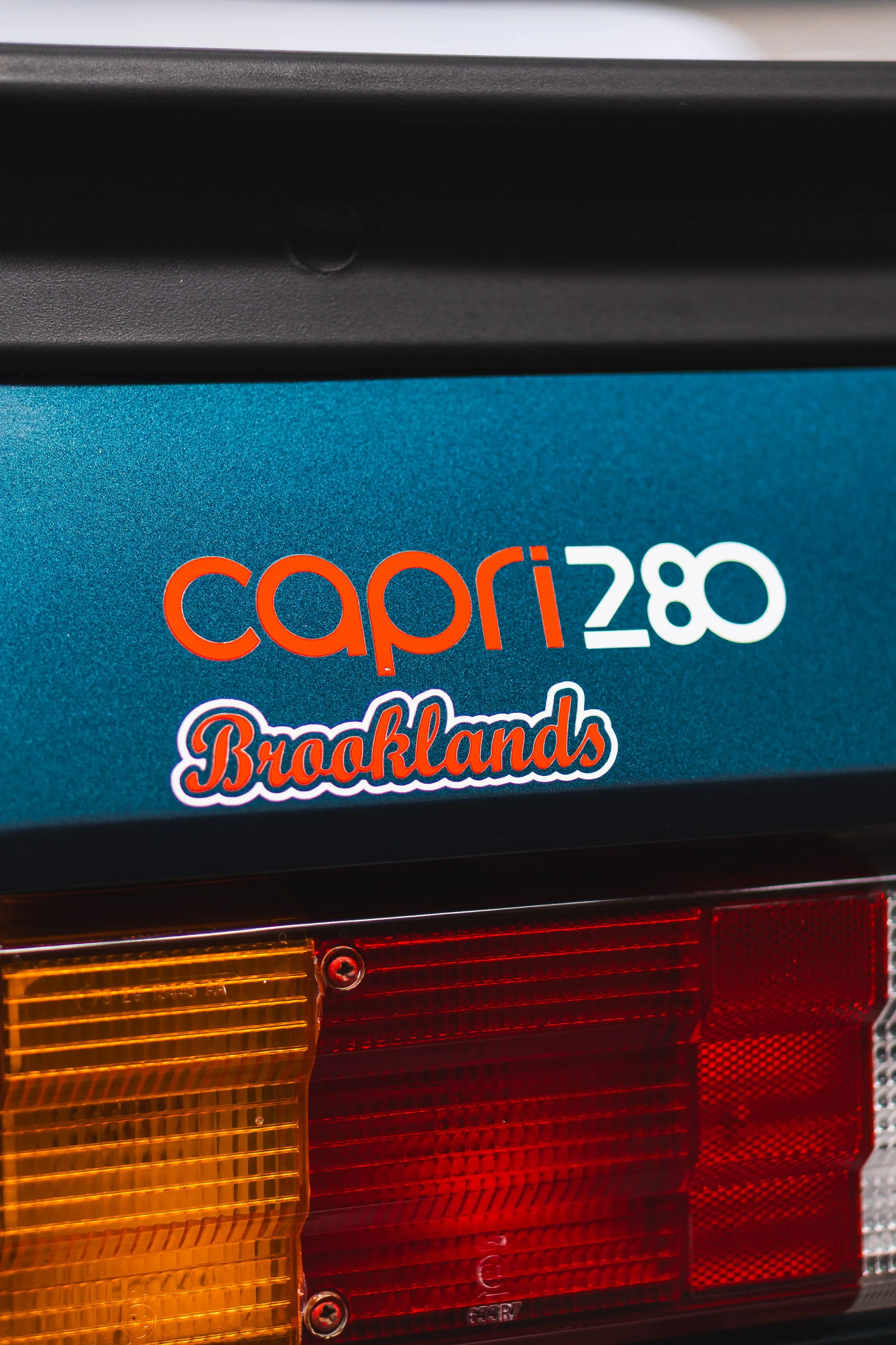 Close-up of the rear of a vehicle showing a sticker that reads 'capri 280' and another sticker that says 'Brooklands,' with part of the taillight visible.