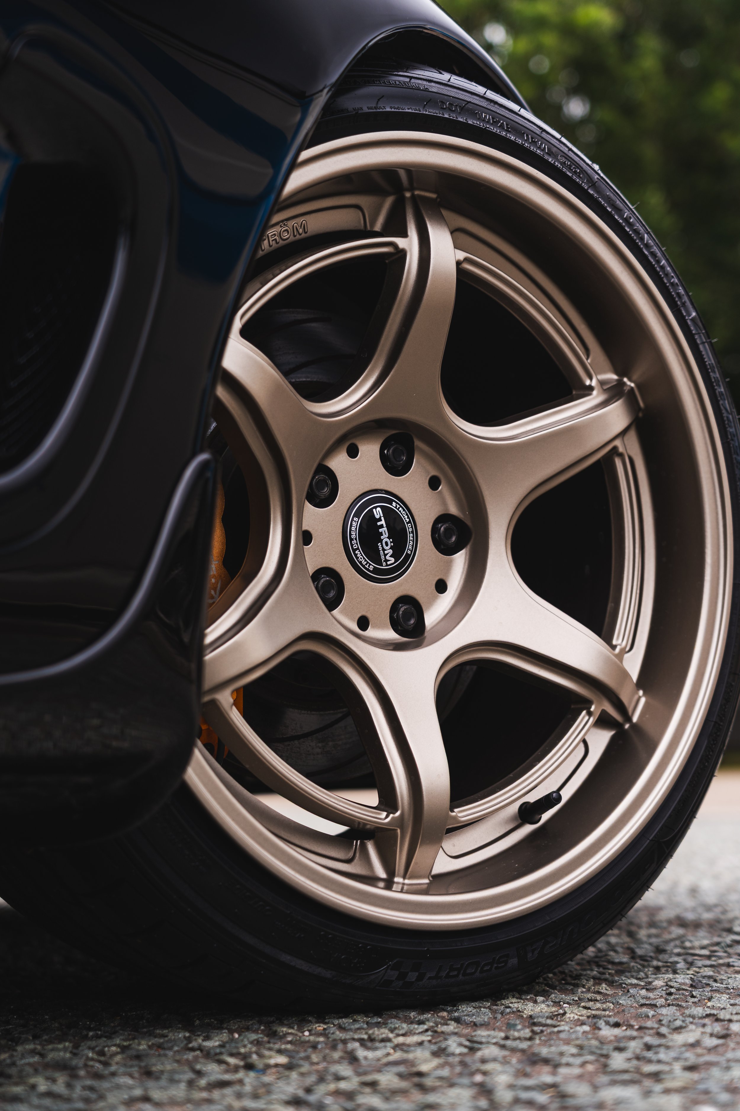 Close-up of a car wheel with a gold-colored rim on a rough asphalt surface, with a blurred green background.