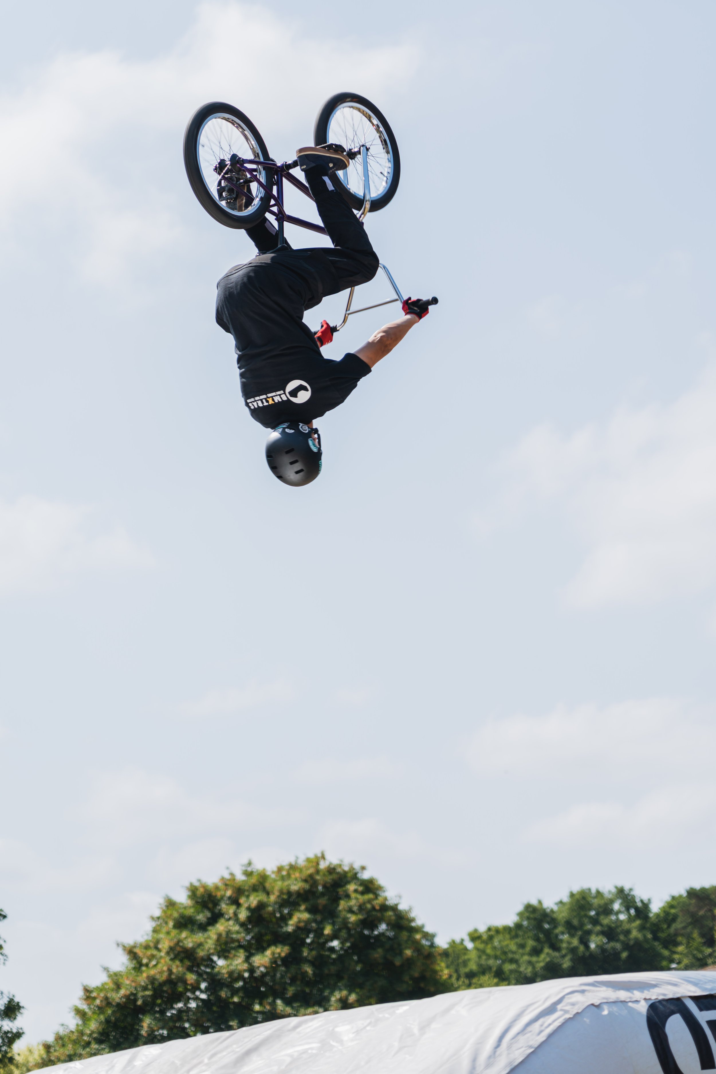 A person performing a stunt on a BMX bike mid-air, upside down, against a cloudy sky and green trees in the background.