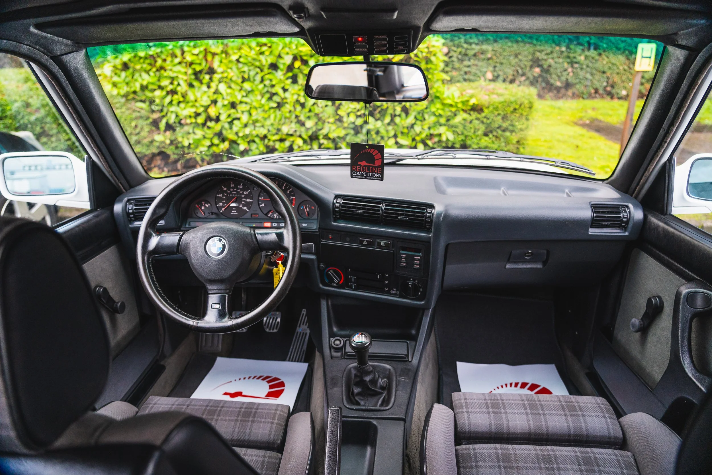Interior view of a car from the driver's seat looking forward, showing the steering wheel, dashboard, rearview mirror, and windshield with greenery outside.