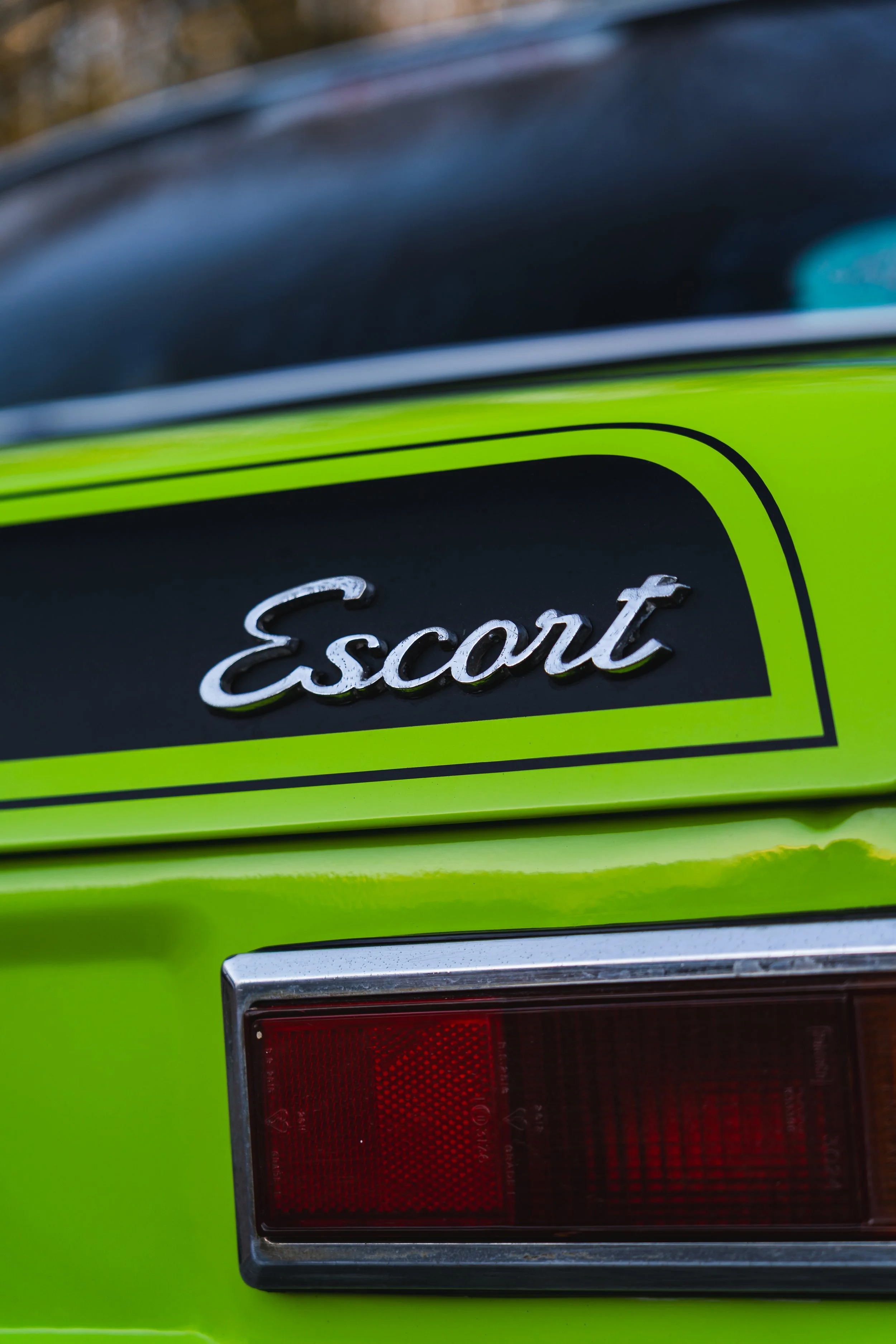 Close-up of the rear of a green vehicle, showing a black badge with the word 'Escort' in silver cursive lettering and a red tail light.
