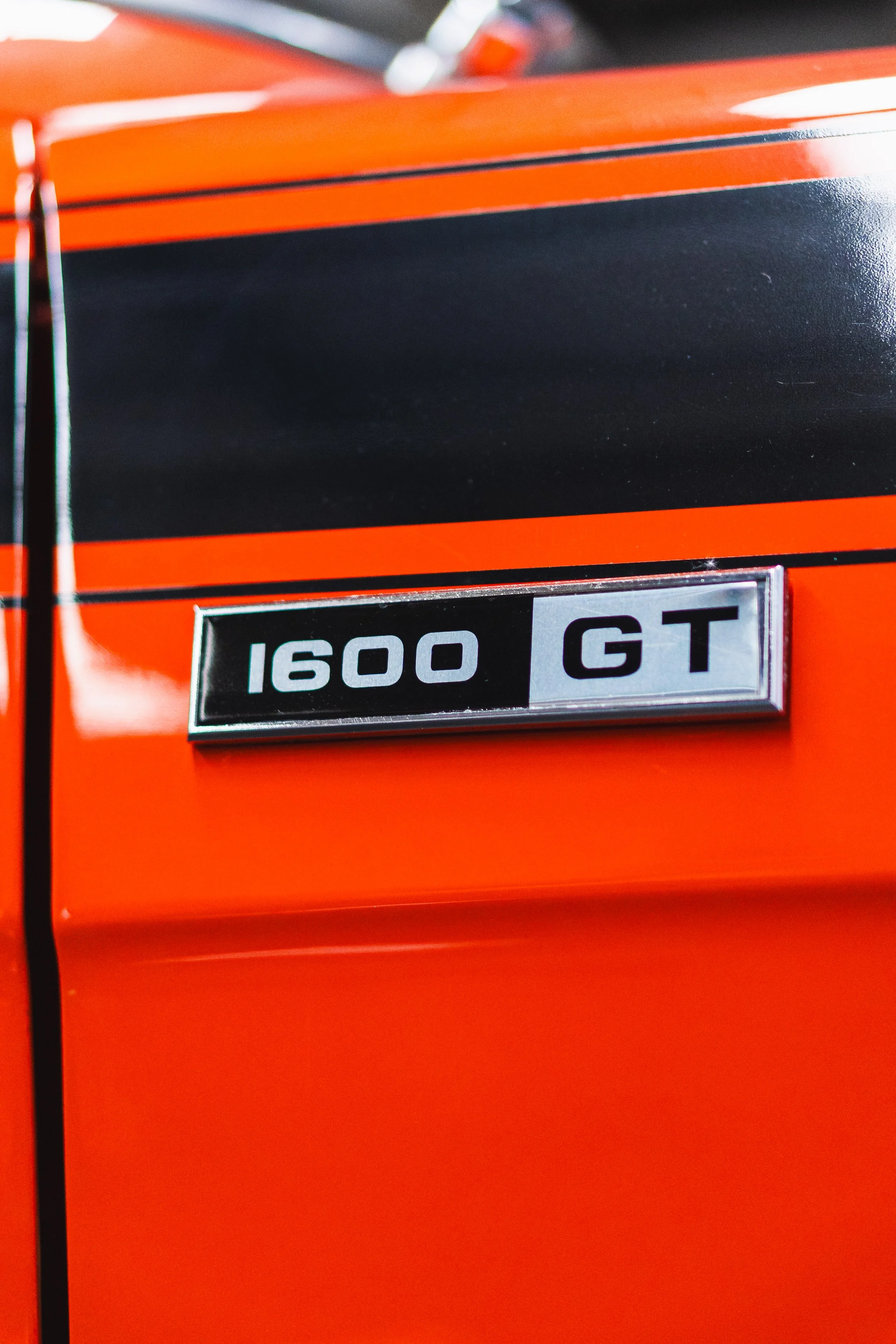 Close-up of a vintage car badge showing '1600 GT' on a bright orange and black car.
