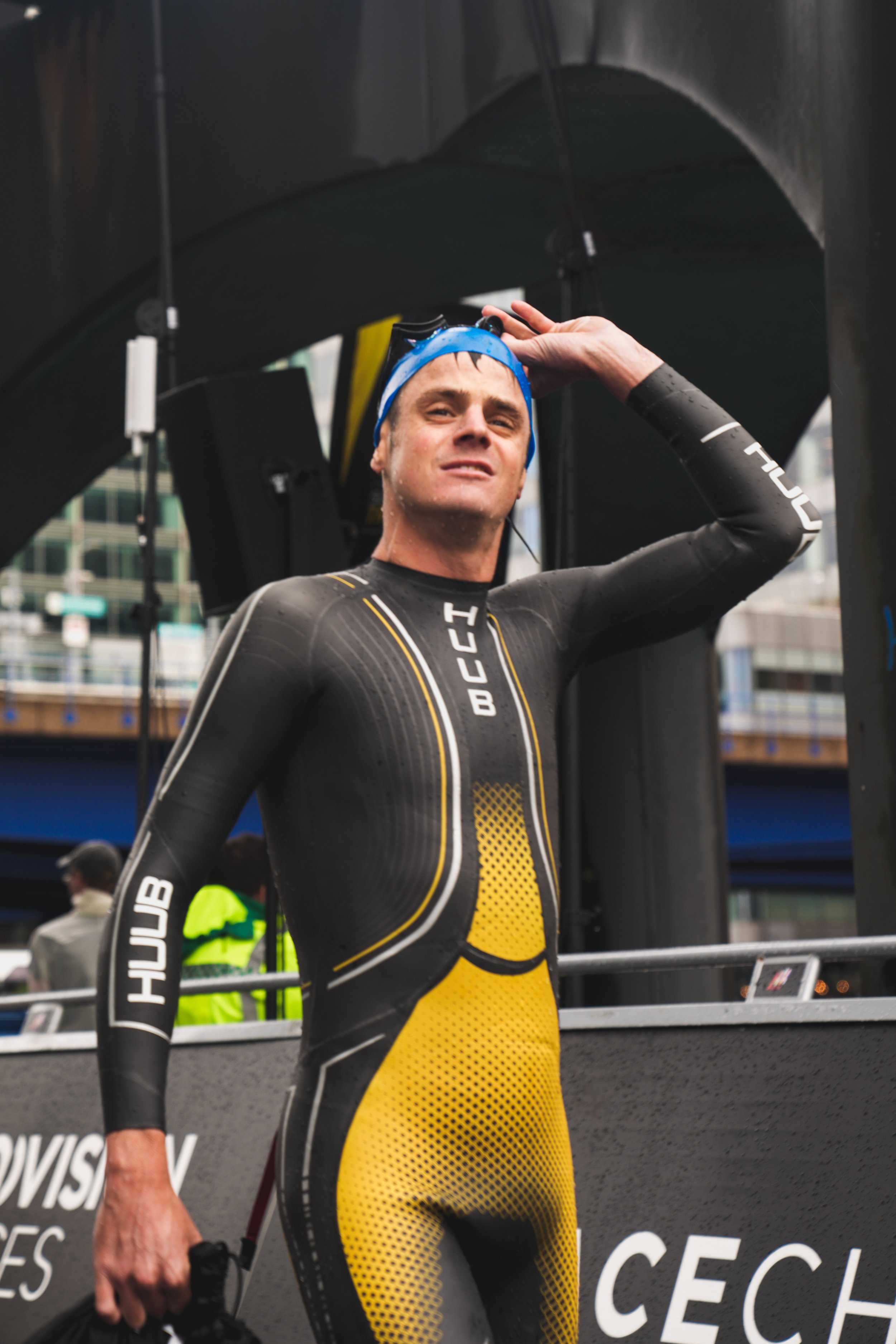 A male triathlete in a black and yellow wetsuit standing with one hand on his head, after completing a race, with a finish line structure and city buildings in the background.