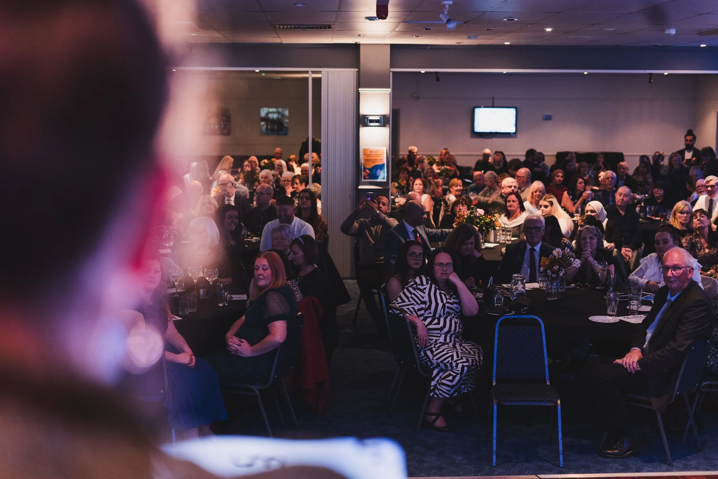 Audience attending a formal event or banquet in a large conference room with decorated tables and floral centerpieces, some people taking photos or looking towards the front.