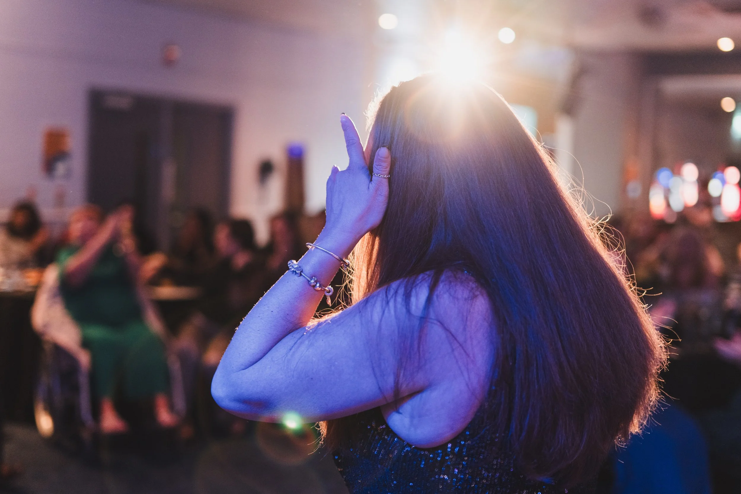 A woman with long hair, wearing jewelry, covering her face with her hand in a dimly lit room with other people sitting at tables and bright lights in the background.