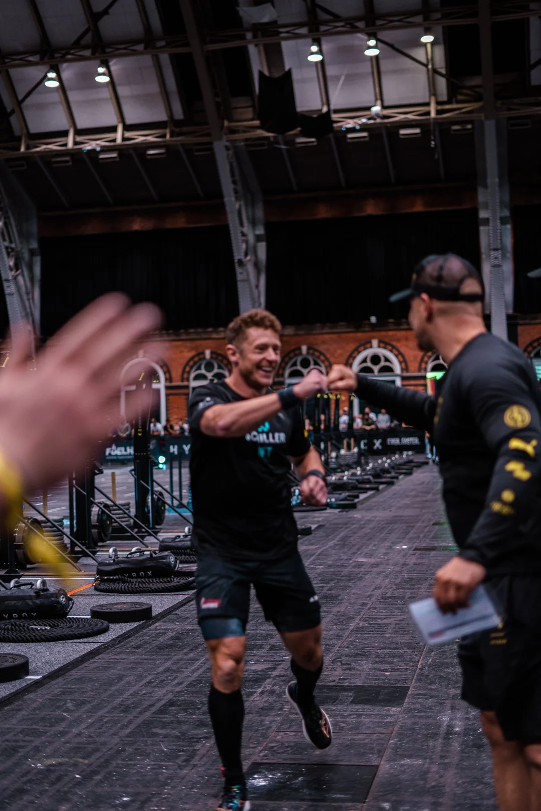 A man in athletic clothing smiling and fist-bumping another person in an indoor gym with workout equipment and a brick wall background.