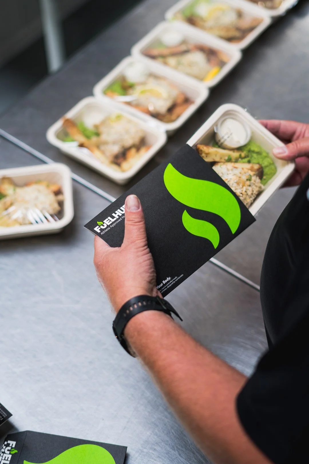 Person holding a boxed meal with a black and green branded sleeve, with several similar meals on a table in the background.