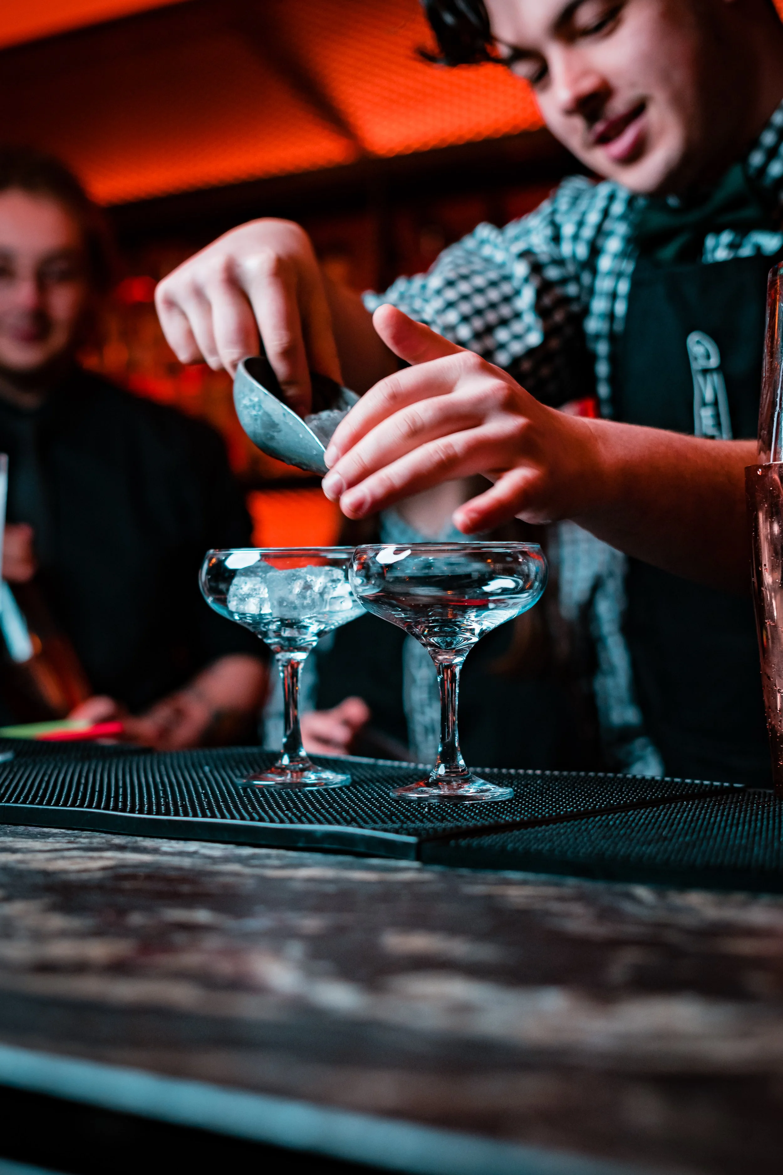 A bartender is pouring a drink into cocktail glasses at a bar with orange lighting, with people watching in the background.