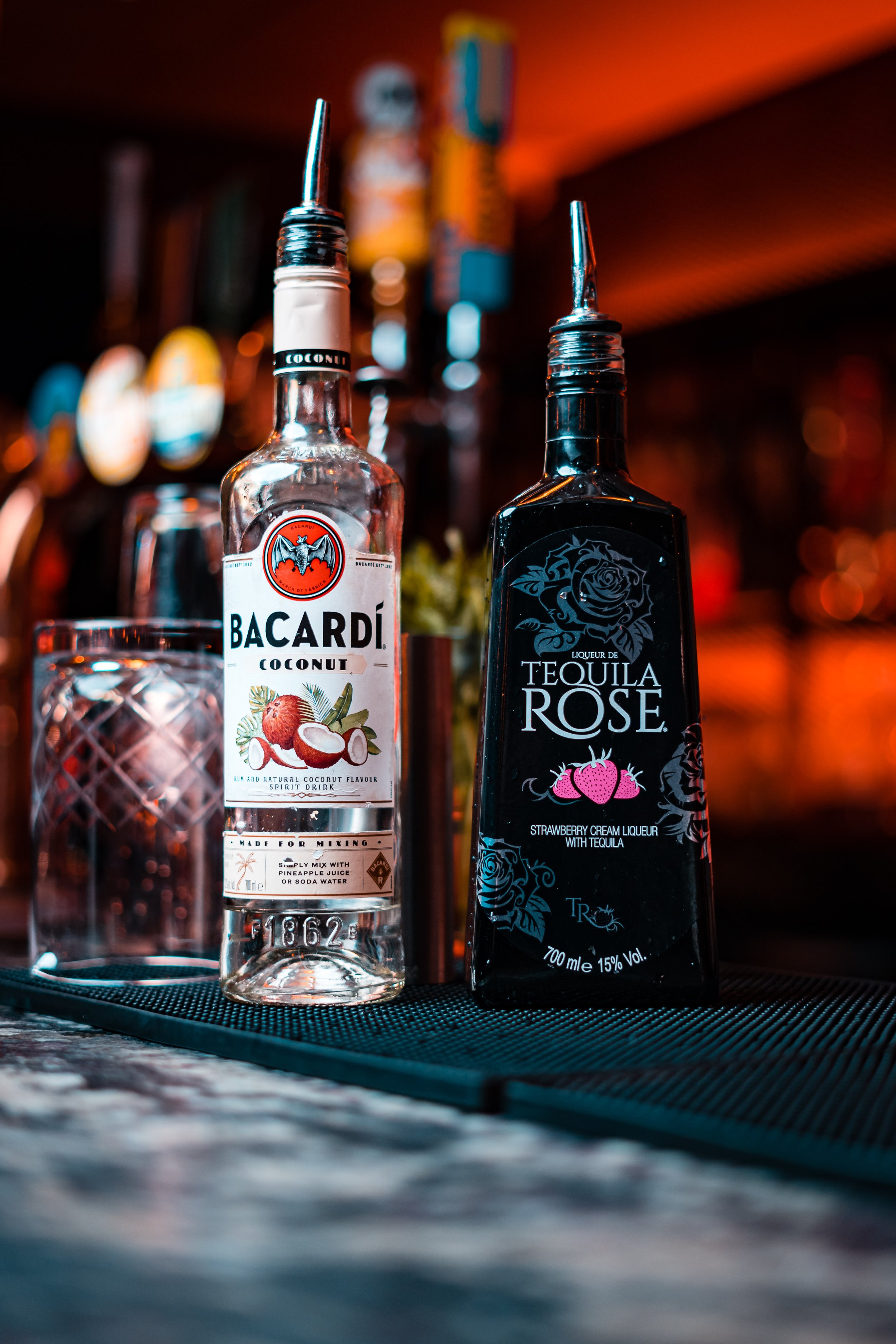 Two bottles of flavored liqueur, Bacardi Coconut and Tequila Rose, with pouring spouts, placed on a bar counter with a glass, against a blurred bar background with warm lighting.