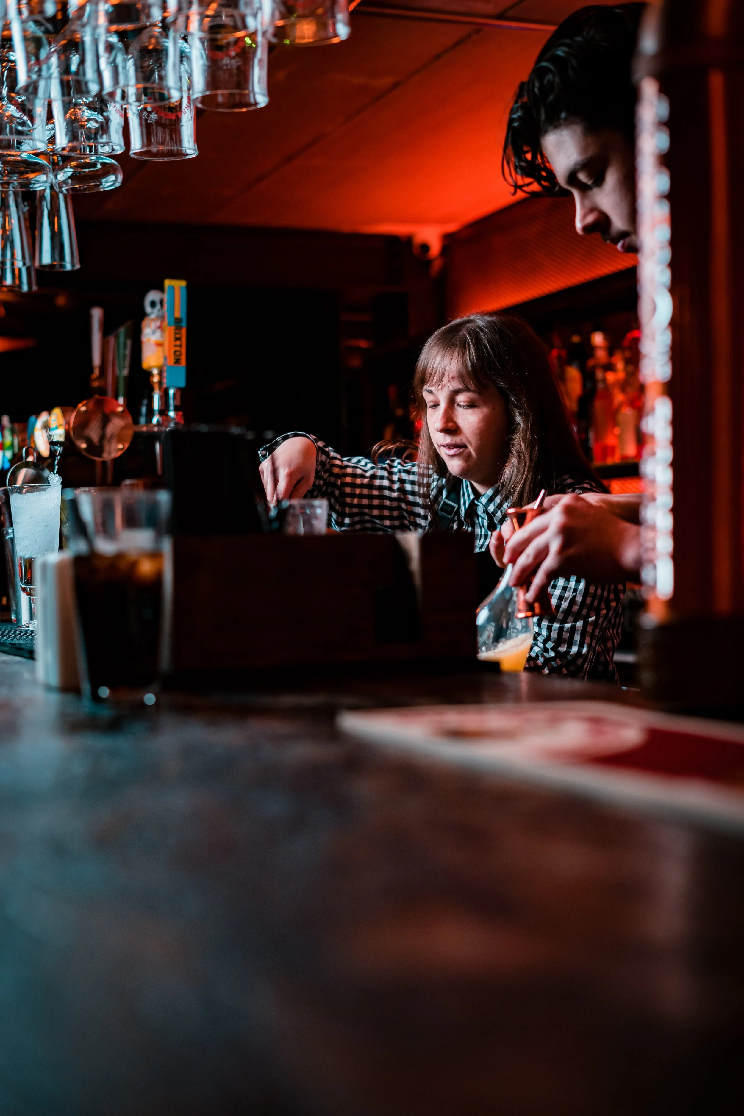 Two bartenders preparing drinks behind a bar at night, with hanging glassware above. One woman with long brown hair is focusing on mixing a drink, and a man with dark, wet hair is also preparing a beverage.