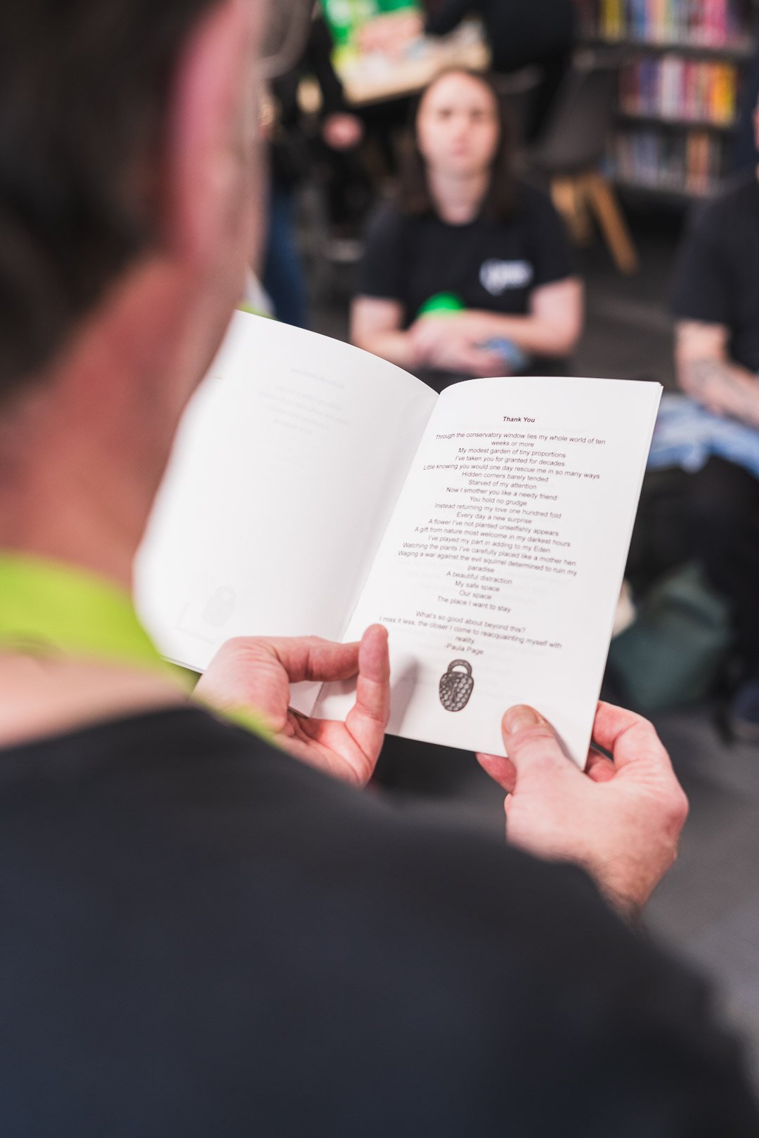 Person holding open booklet with text and small drawing of a matchstick on the pages, others sitting in background at what appears to be a bookstore or library.