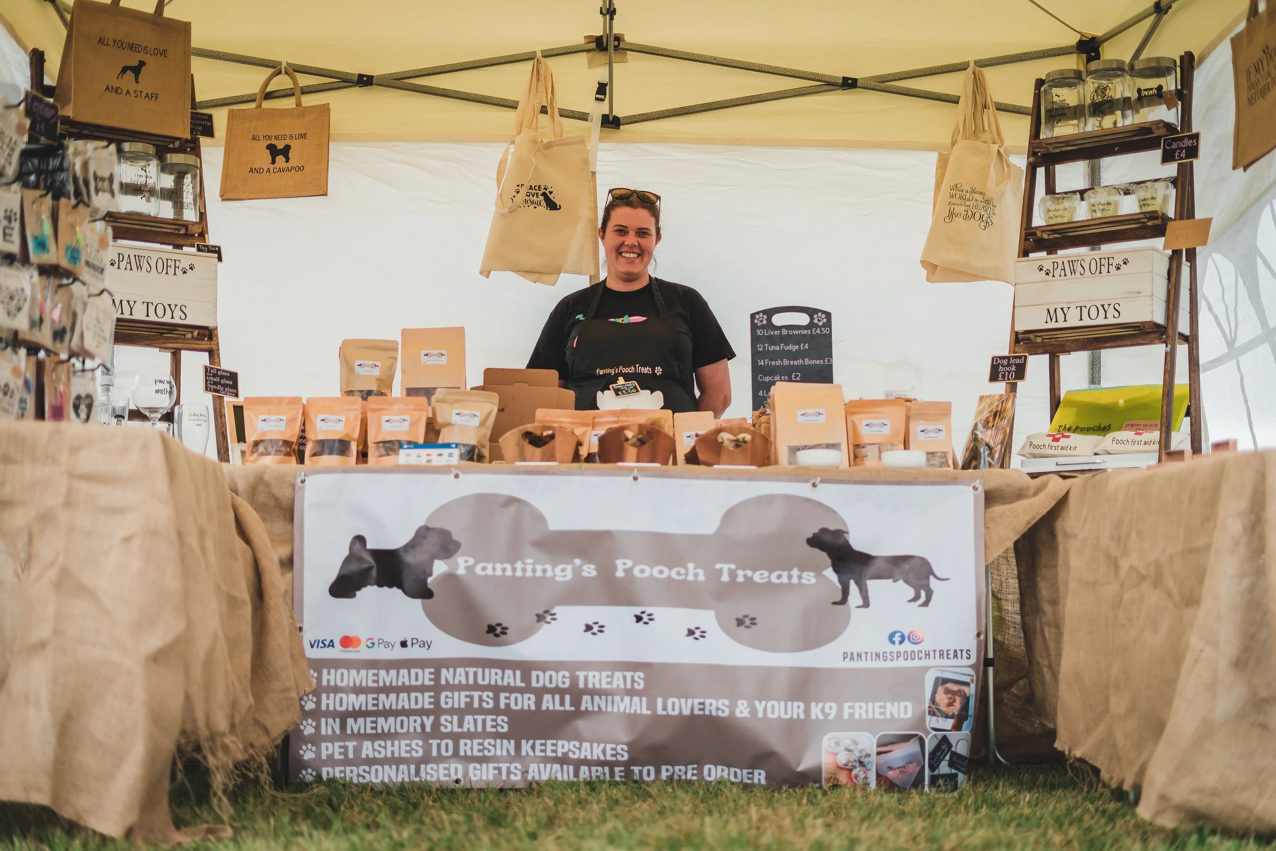 A woman stands behind a stall selling homemade dog treats, with signs and bags displaying the products at an outdoor market.