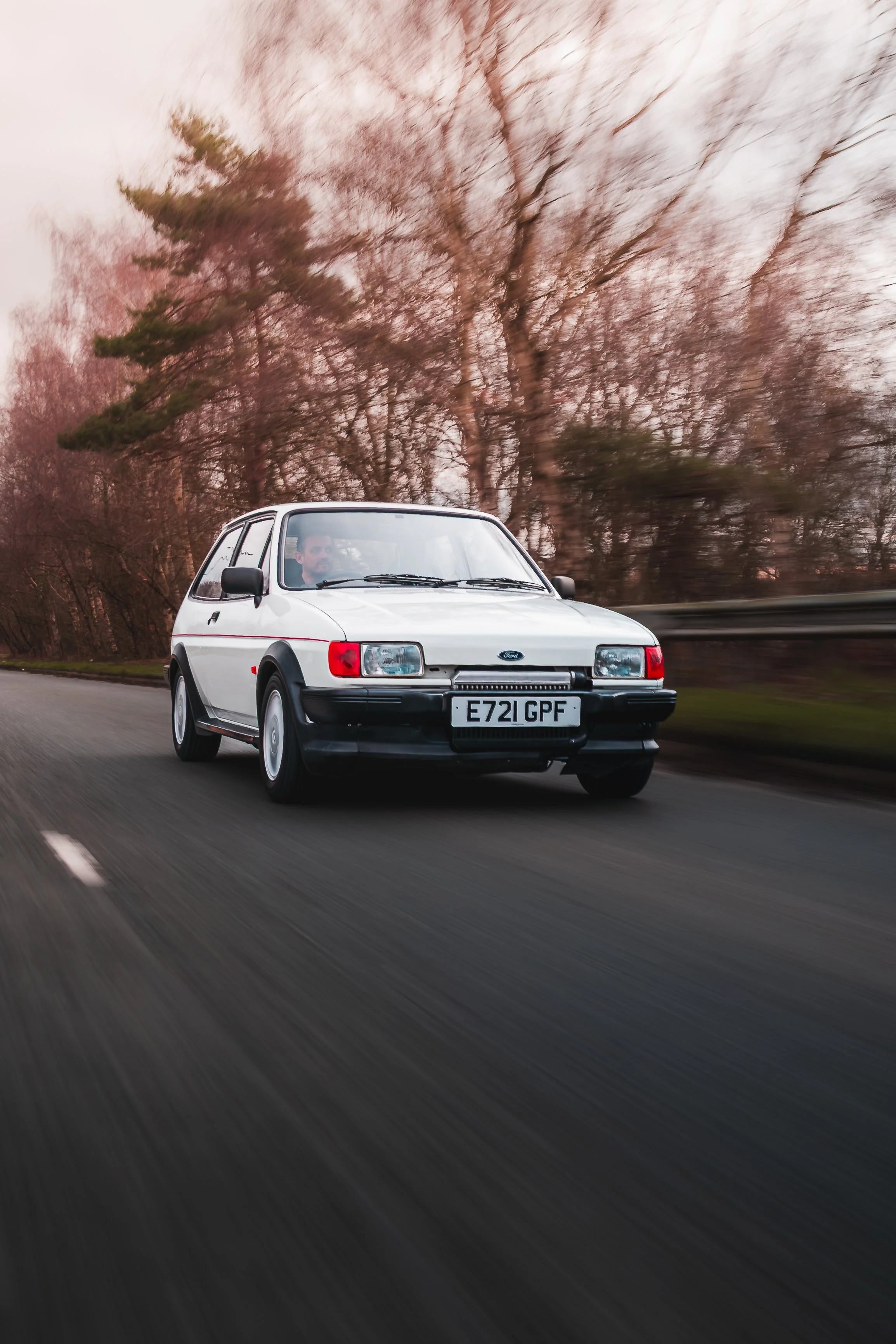A white vintage Ford car driving on a road with trees in the background.