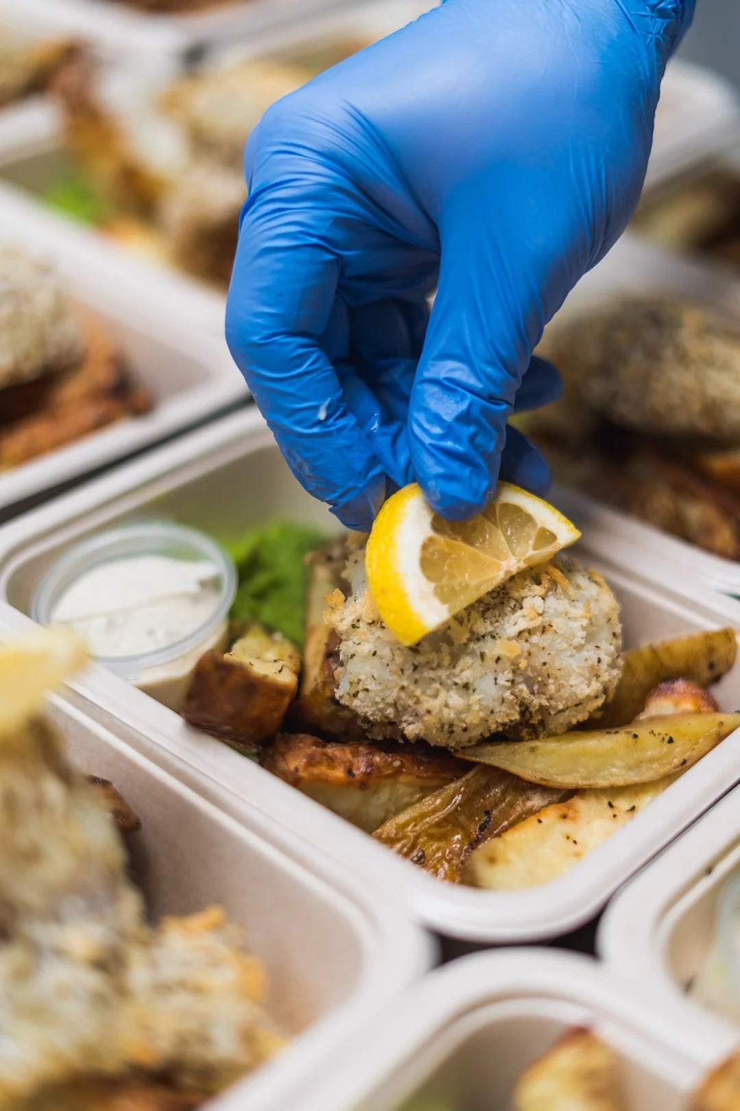A hand wearing a blue glove is placing a lemon wedge on a breaded fish cake in a takeout container with fried potato wedges and a small cup of dipping sauce.