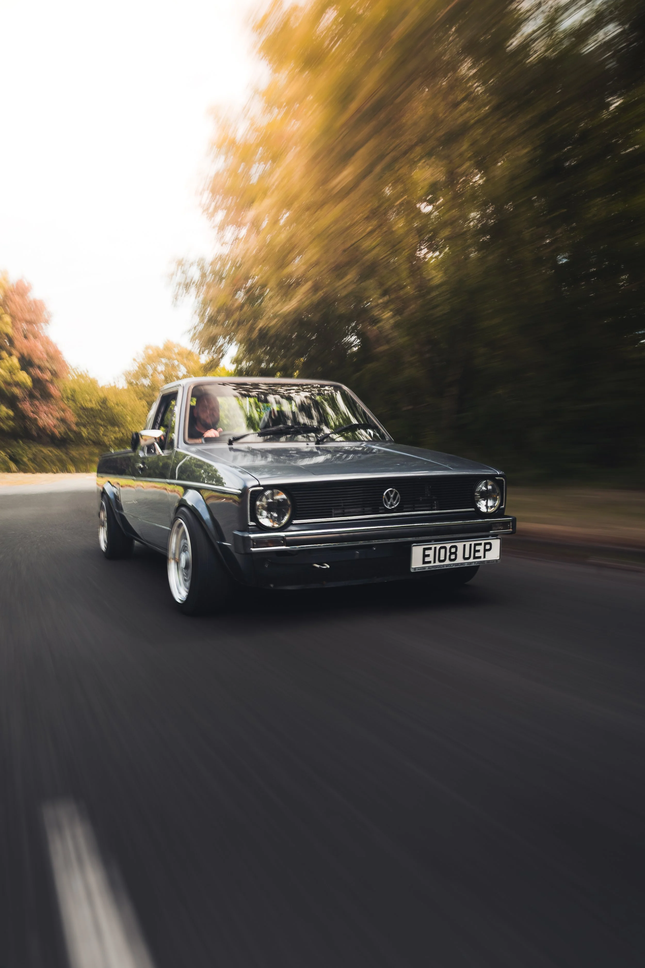 A black vintage Volkswagen car driving on a road surrounded by trees with autumn foliage, motion blurred to indicate speed.