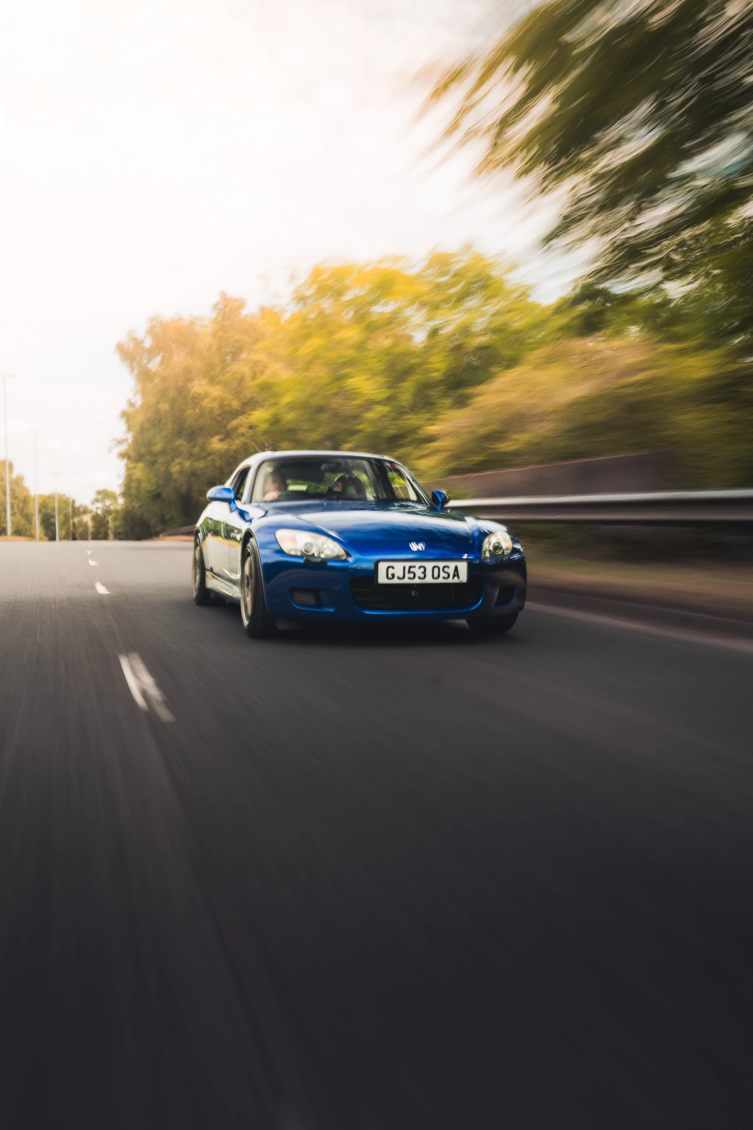 A blue sports car with a license plate GJ53 OSA is driving on a highway surrounded by trees with blurred leaves due to speed.