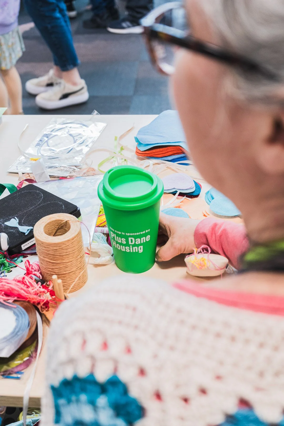 A person sitting at a table with various craft supplies, including yarn, fabric, and paper cutouts, holding a green tumbler that says 'Sponsored by Plus Dane Housing.' There are people in sneakers walking in the background.