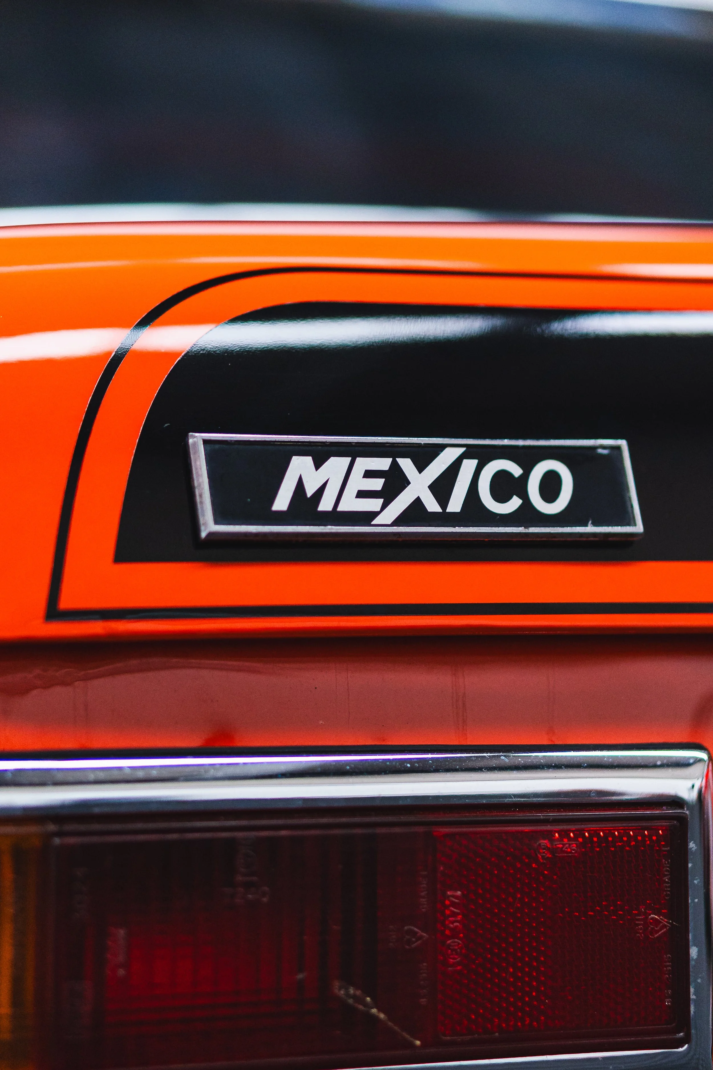 Close-up of a vintage car's rear, showing a black and orange paint job and a badge that reads 'MEXICO'.