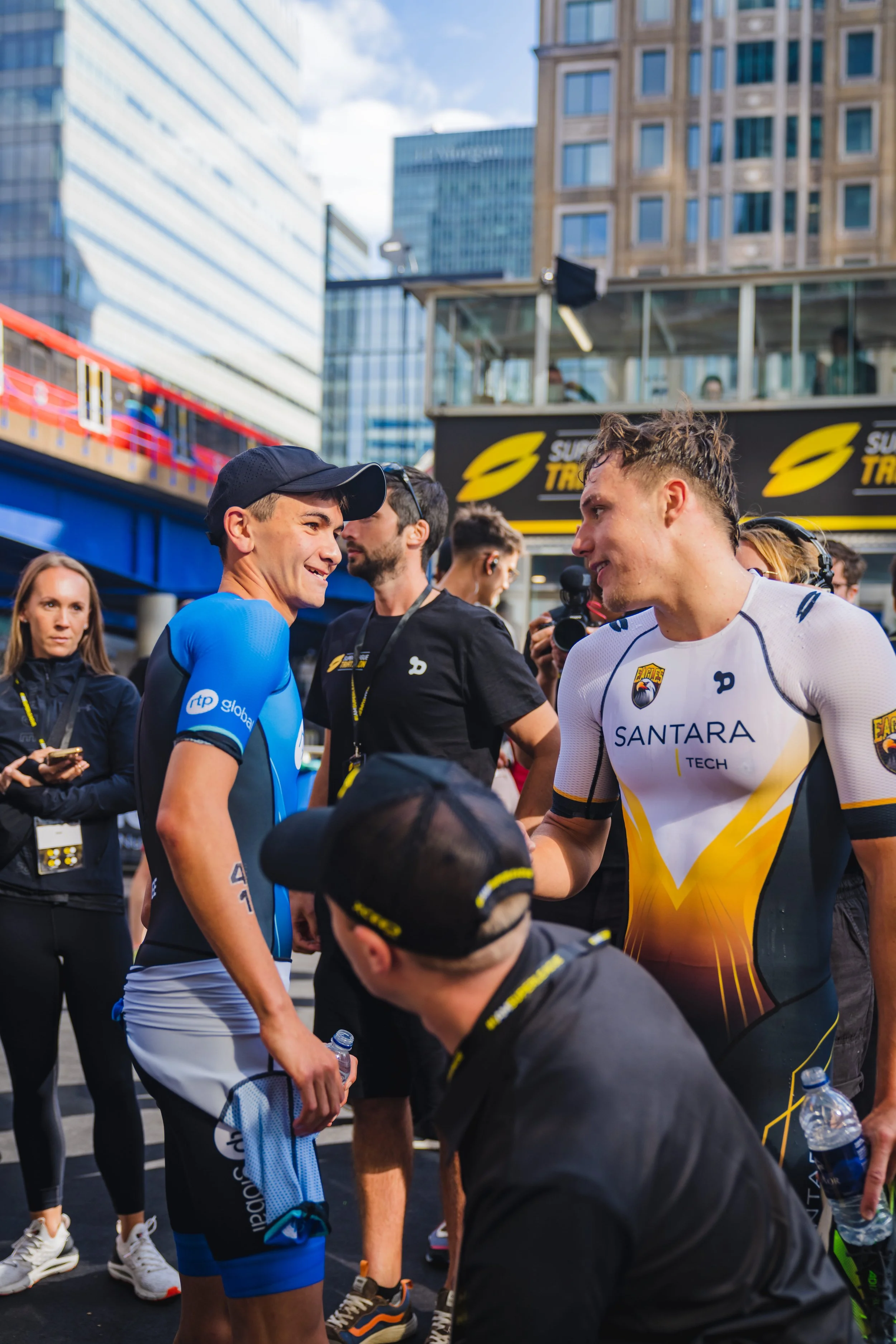 Two male athletes, one in a blue sports uniform and the other in a white and yellow sports uniform, shaking hands at a race event in an outdoor city setting with tall buildings and a digital display in the background. Several other people and photogr