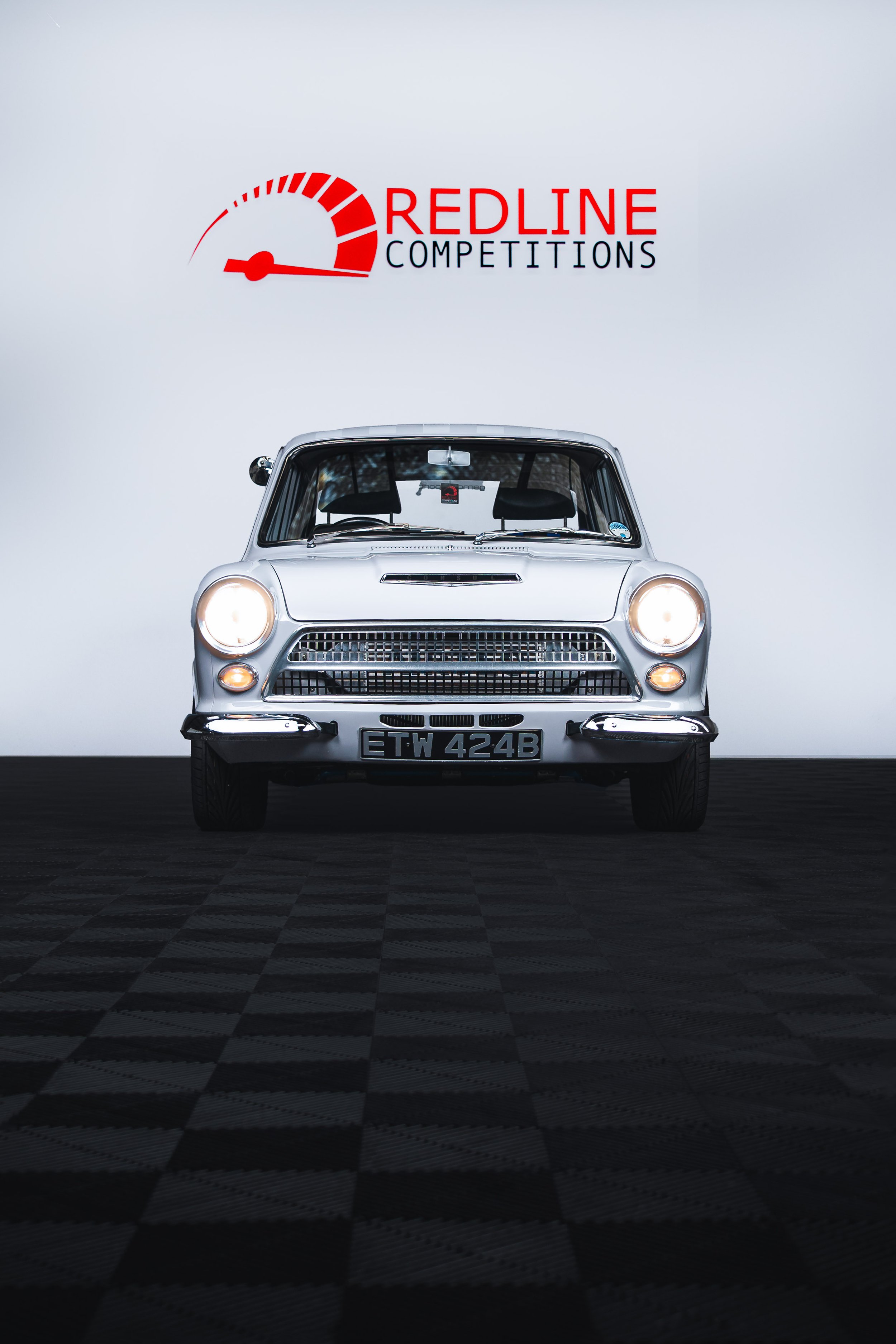 A classic silver car with headlights on, parked in front of a white wall with a "Redline Competitions" logo and a speedometer graphic.
