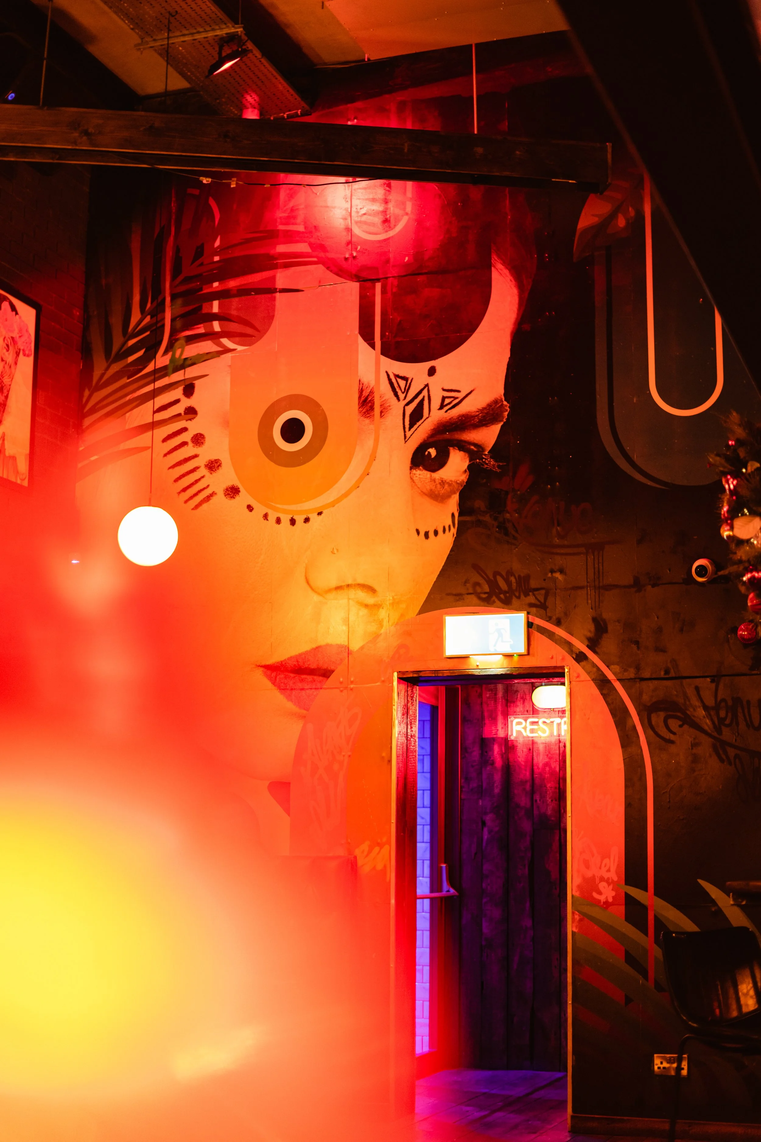 A large mural of a woman's face with colorful face paint and tattoos covers the wall inside a vibrant, graffiti-covered bar or nightclub, illuminated with red and purple neon lights.