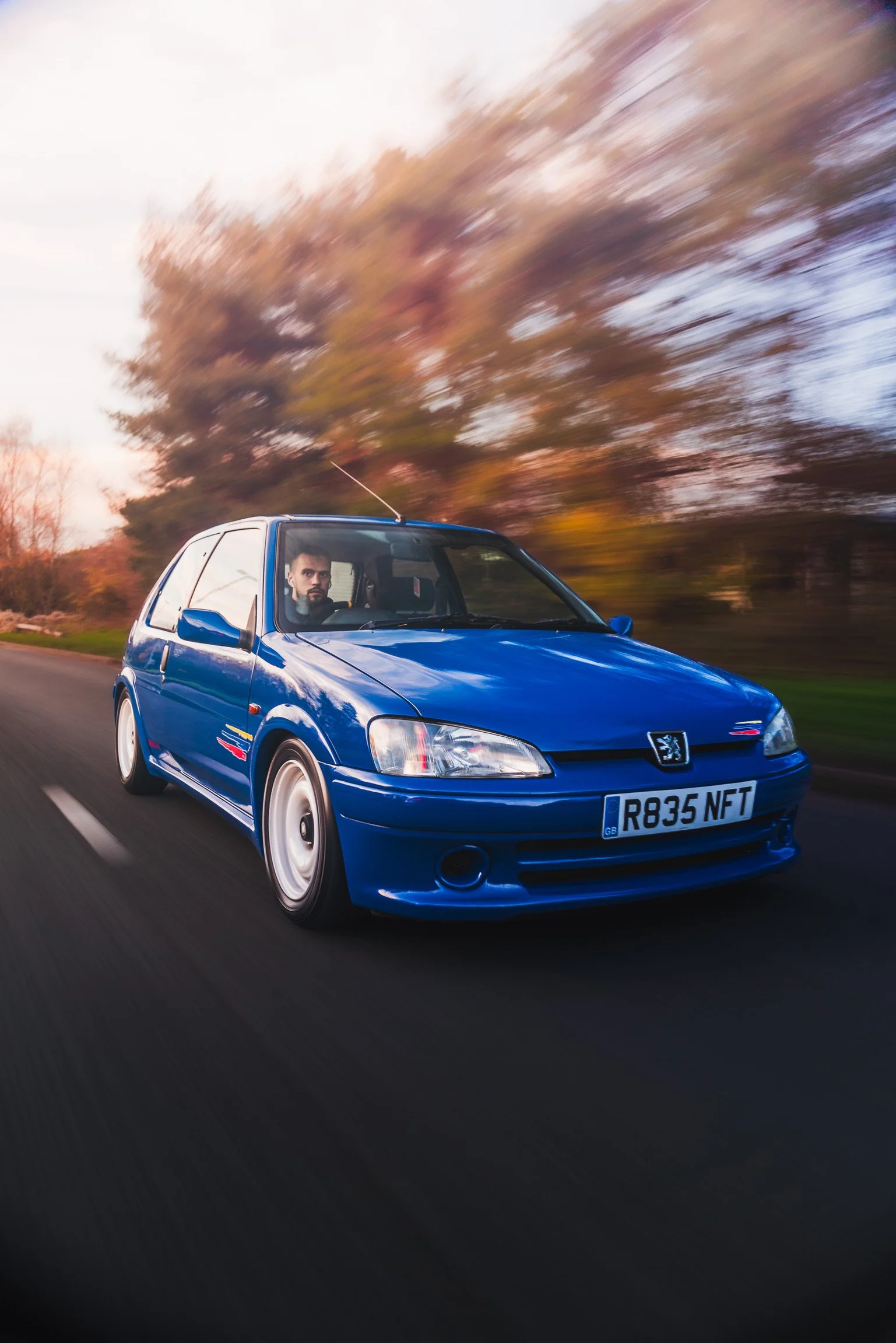 A blue vintage Peugeot car driving down a road with autumn trees in the background during sunset.