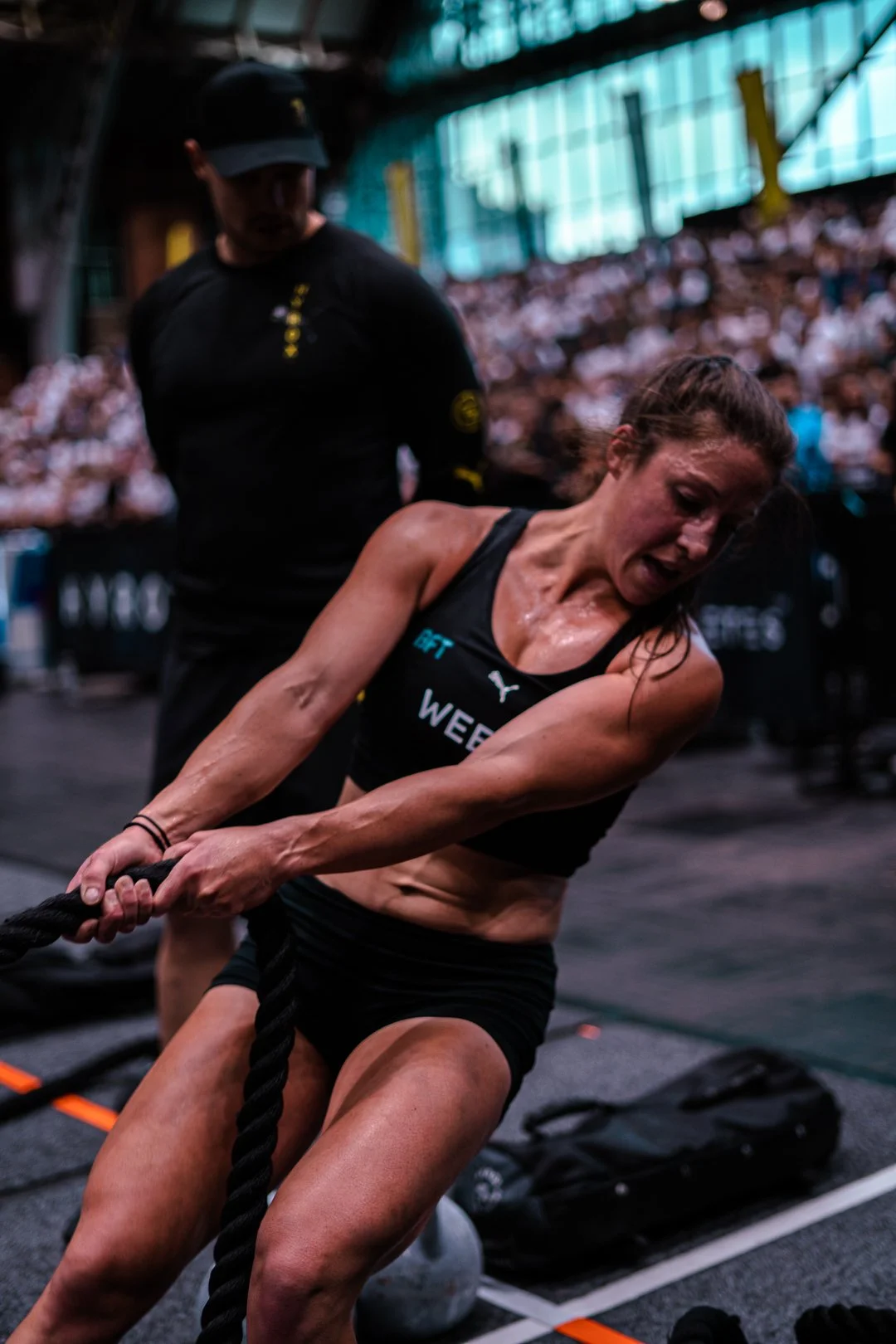 Female athlete during a CrossFit competition pulling a heavy rope, with a coach or trainer behind her and a large audience in the background.