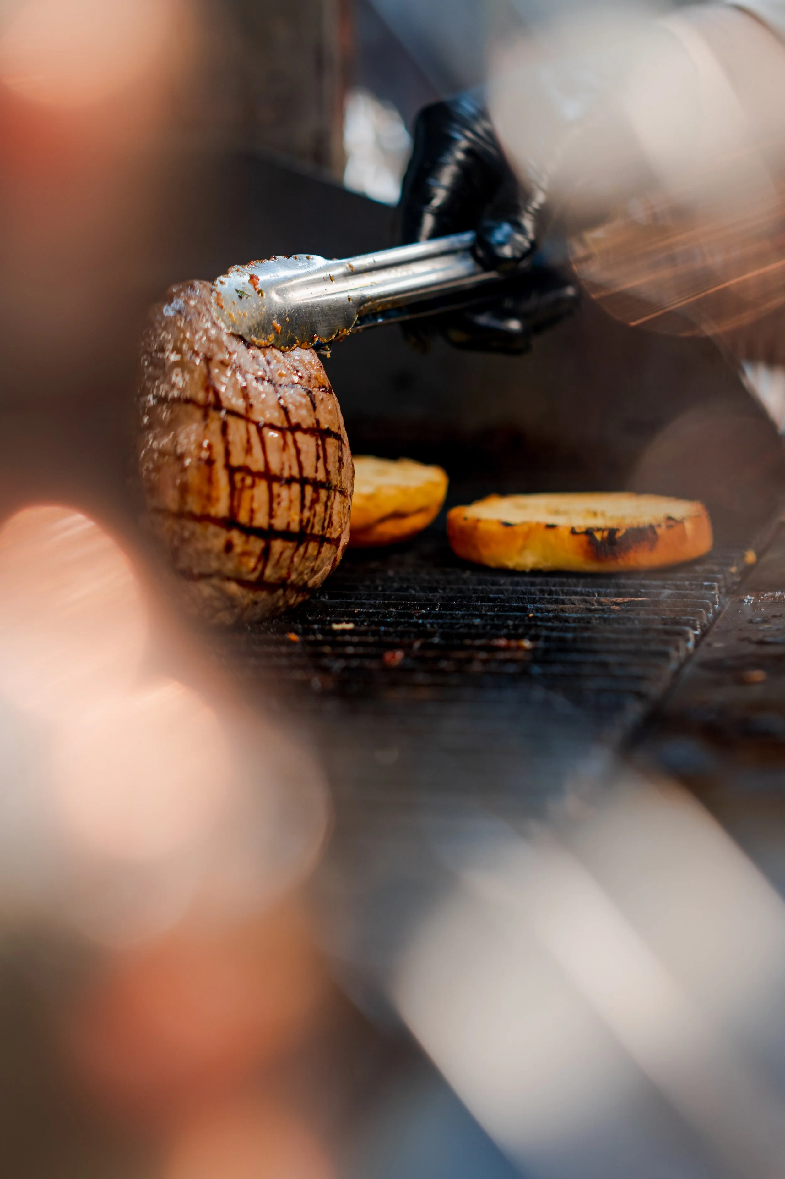 A chef wearing black gloves is grilling a piece of seasoned steak with grill marks, while two grilled banana slices are nearby on the grill.