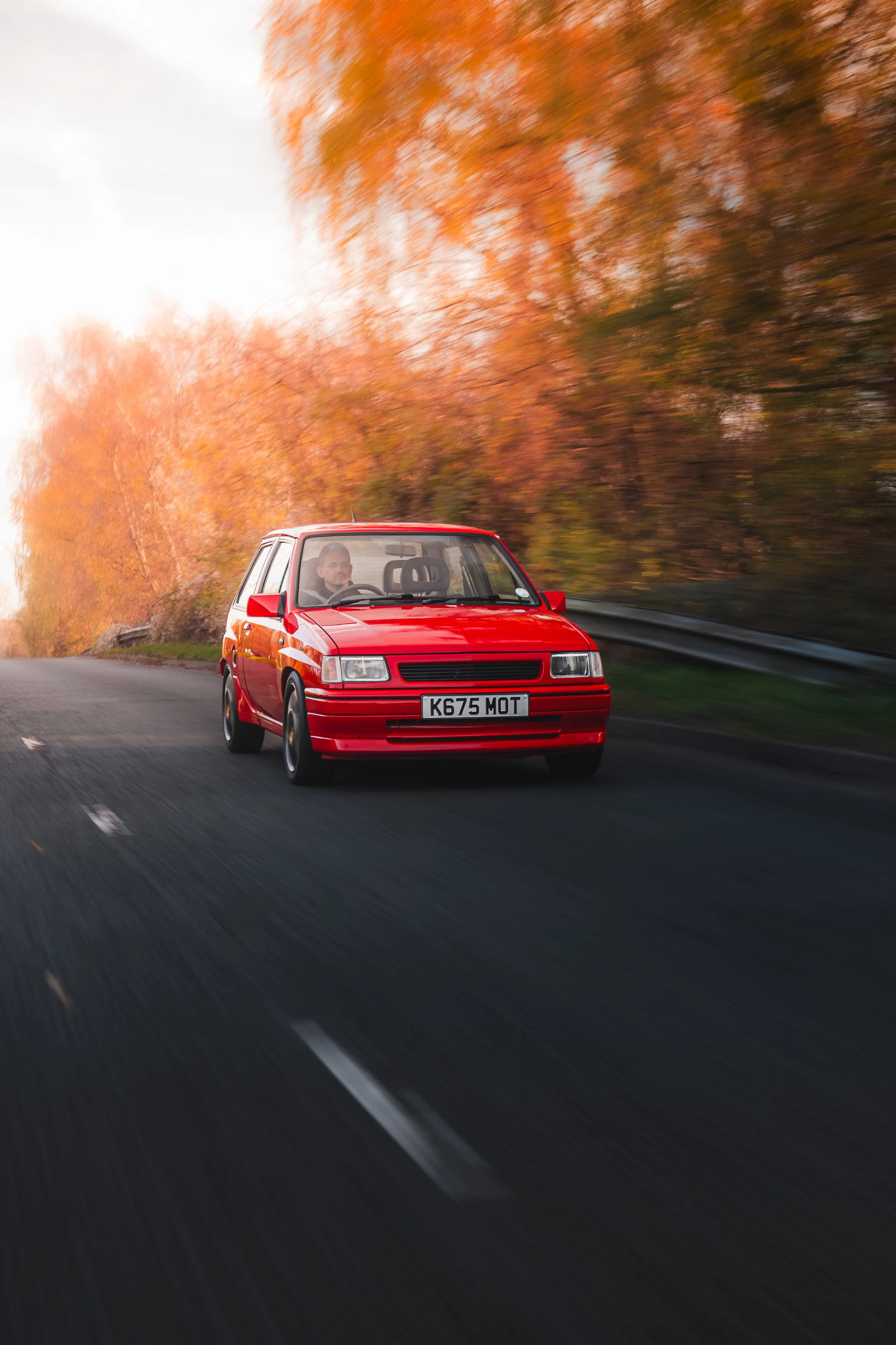 A red compact car driving on a two-lane road with autumn foliage in the background.