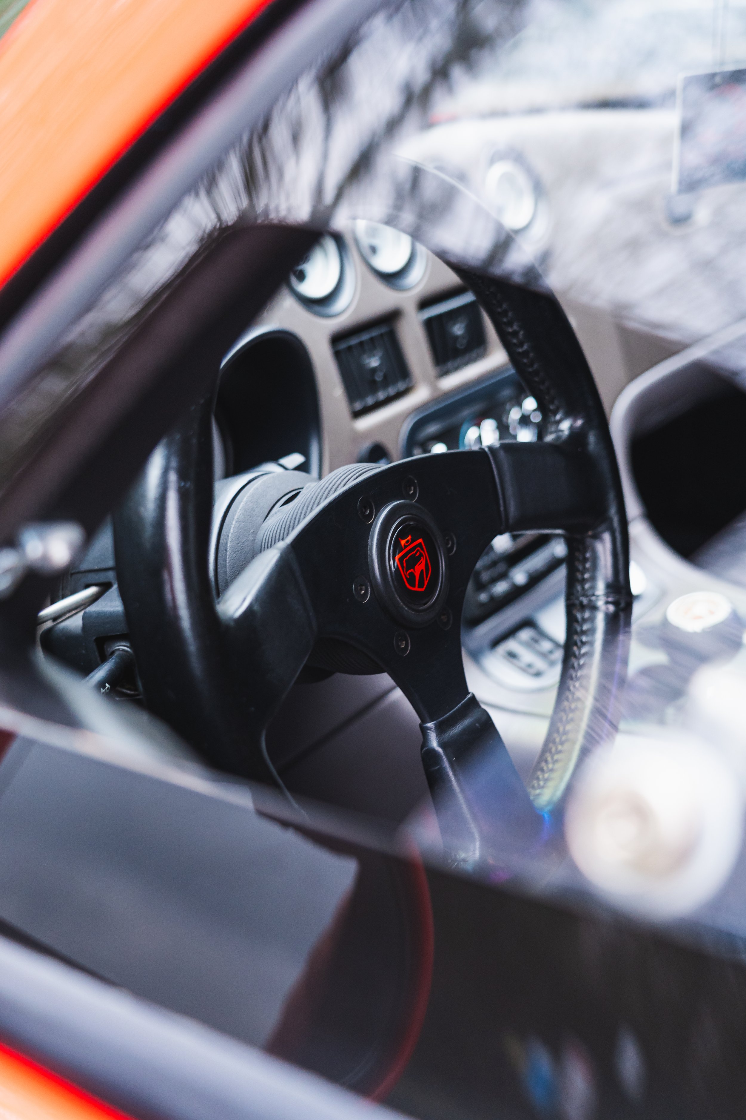 Close-up view of a vintage race car dashboard with a black steering wheel featuring a red emblem at the center, various dials, gauges, and switches visible in the background.