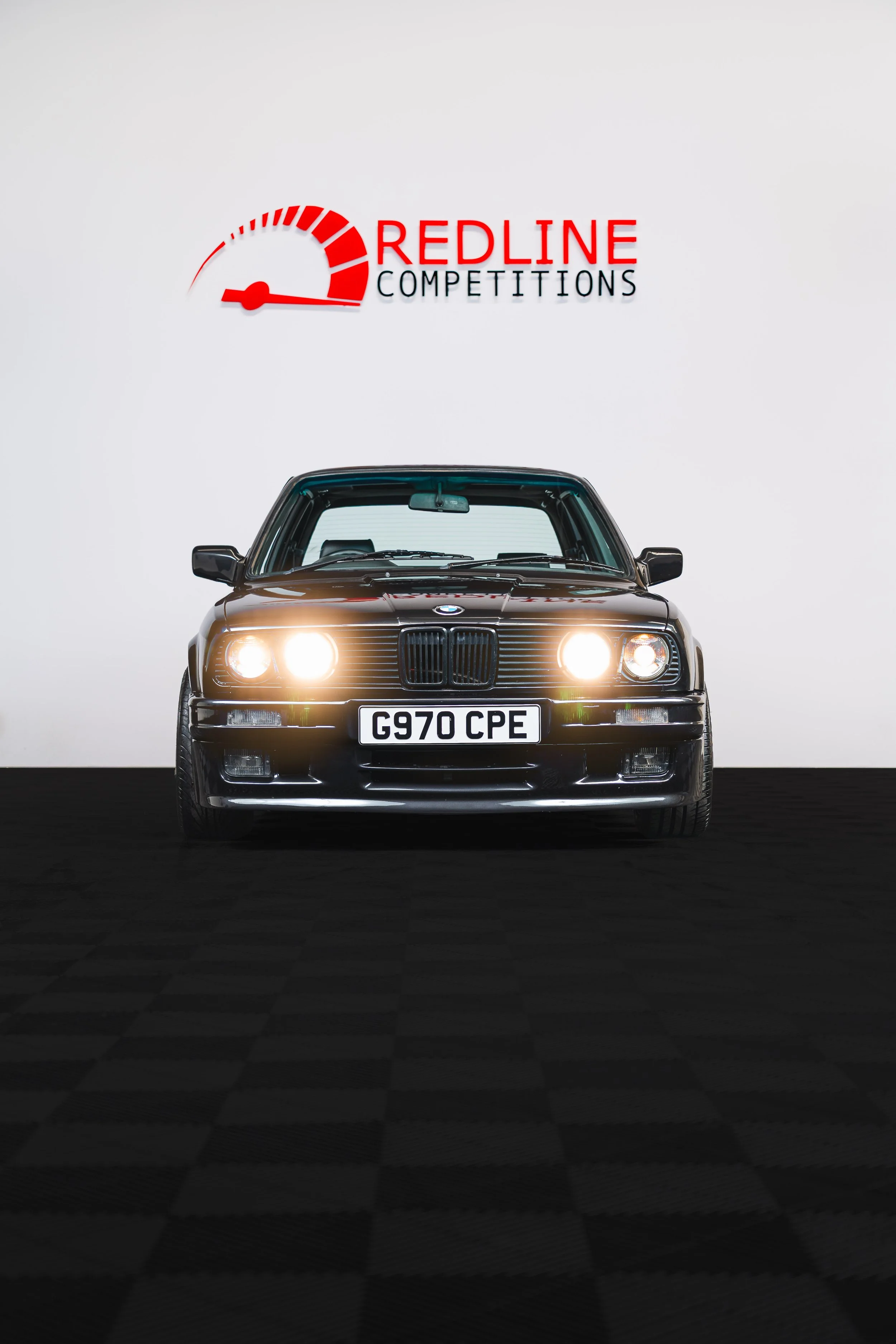 Black vintage BMW car with headlights on, positioned in front of a white wall with a sign that reads 'Redline Competitions' and features a red speedometer graphic.