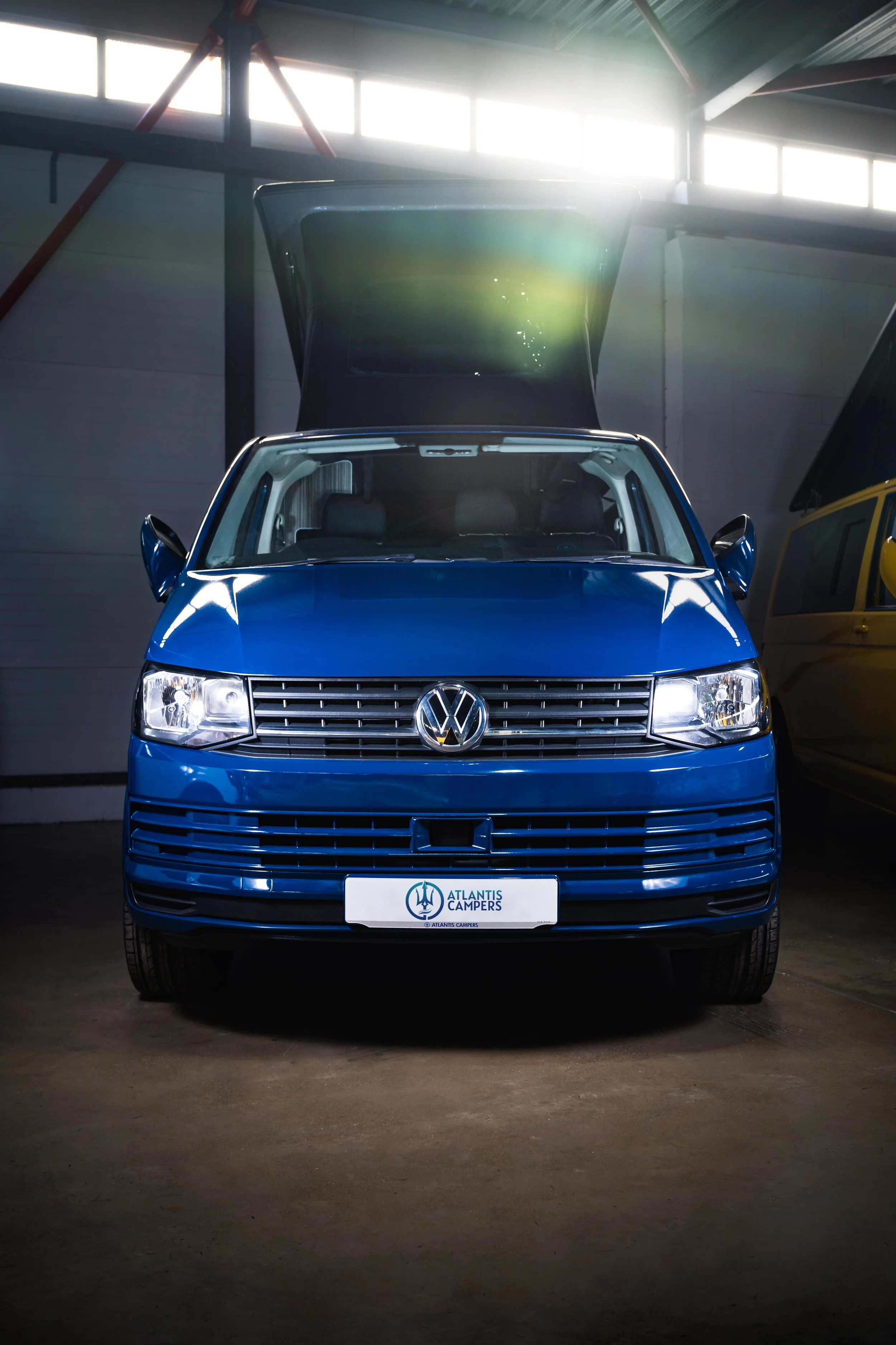 A blue Volkswagen camper van with its rear hatch open inside an indoor garage.