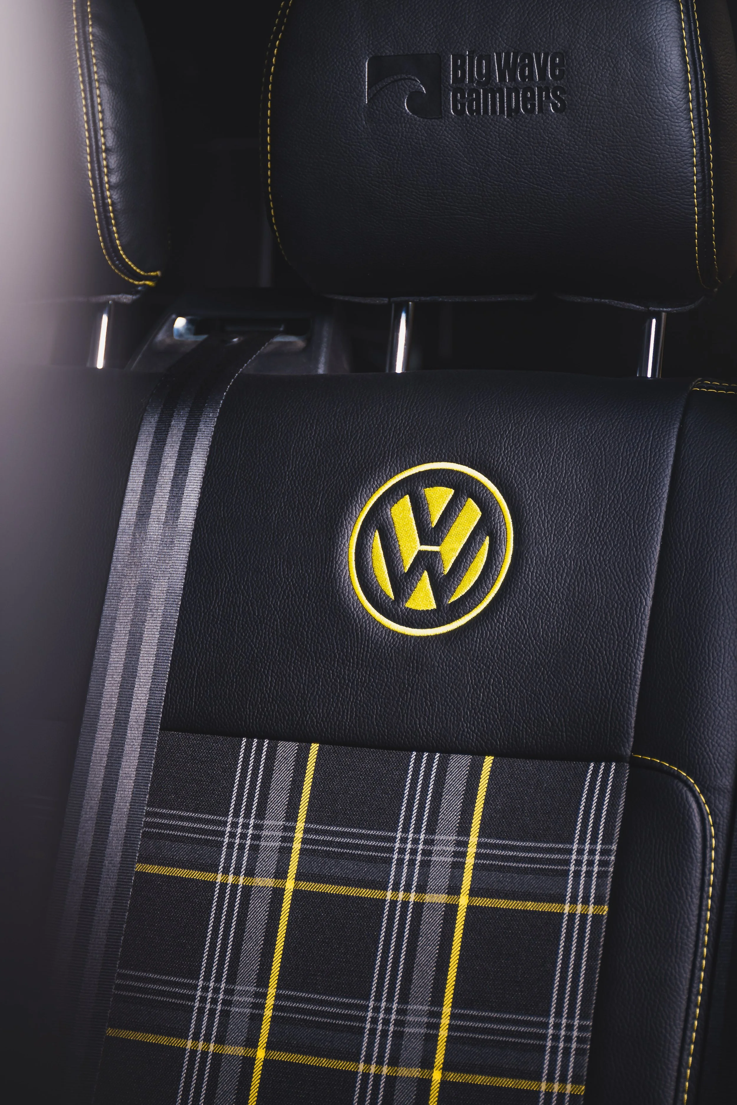 The back seat of a Volkswagen car with a yellow VW logo embroidered on the headrest and yellow stitching on the leather upholstery, complemented by a black and yellow plaid seat cover.