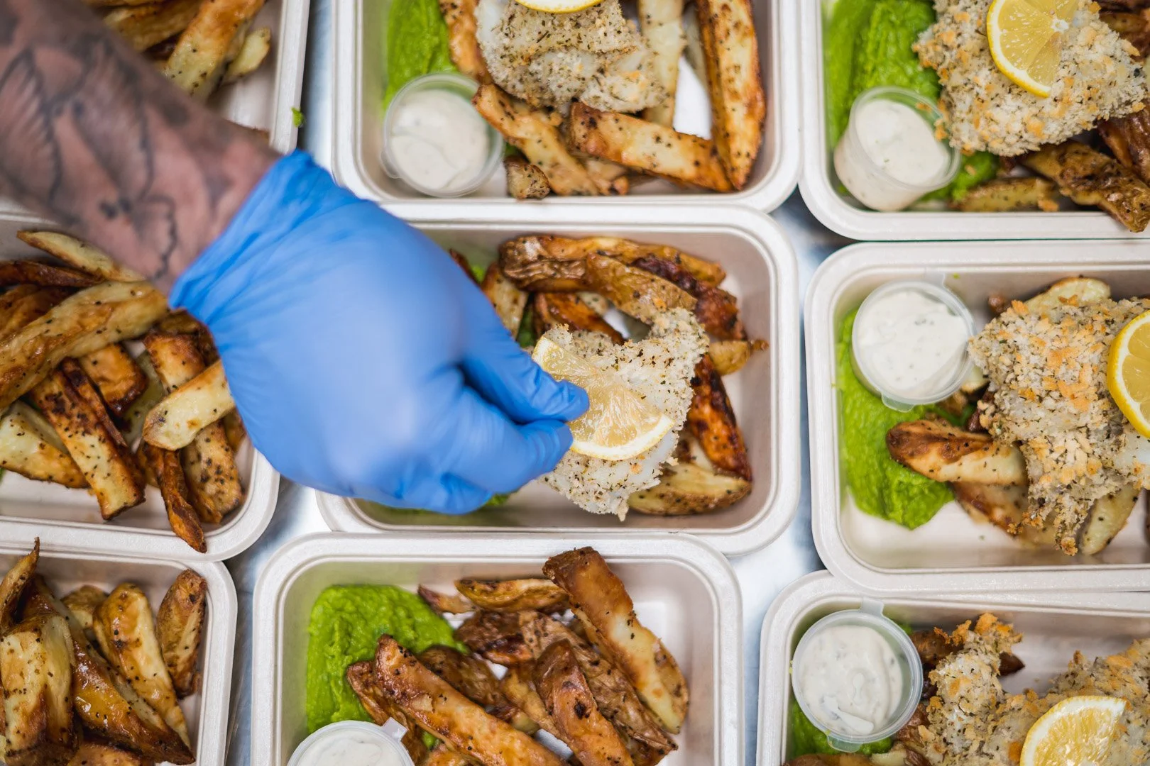 Multiple styrofoam containers filled with fried chicken wings, potato wedges, green lettuce, lemon slices, and small cups of ranch dressing. A hand wearing a blue glove is placing a lemon slice onto one of the containers.